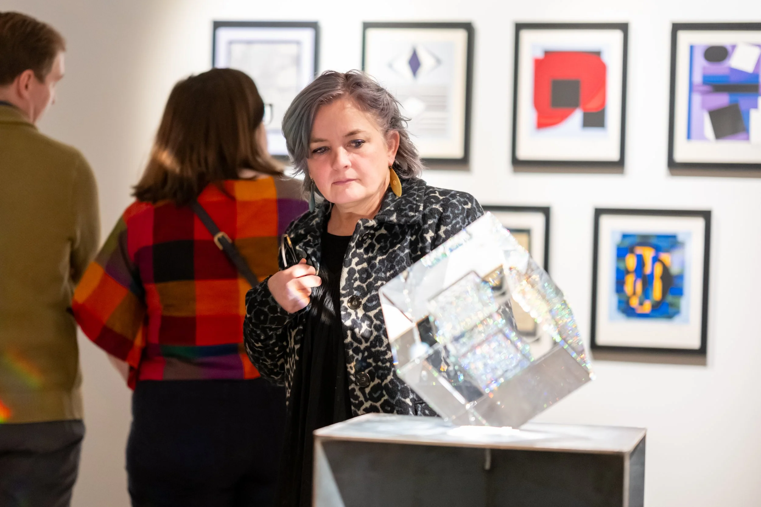 Three people looking at artwork in an art gallery. One woman in a leopard print jacket holding a holographic gift box, with framed abstract art on the wall behind them.