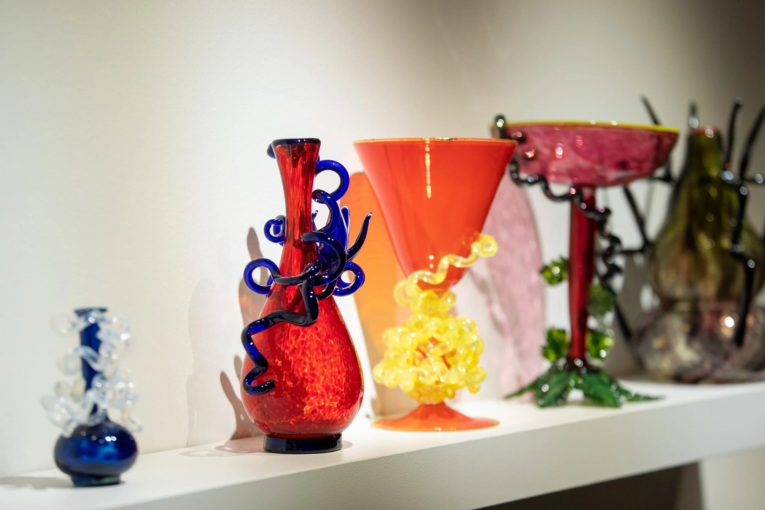 Colorful glass art vases and sculptures displayed on a white shelf against a plain white wall.