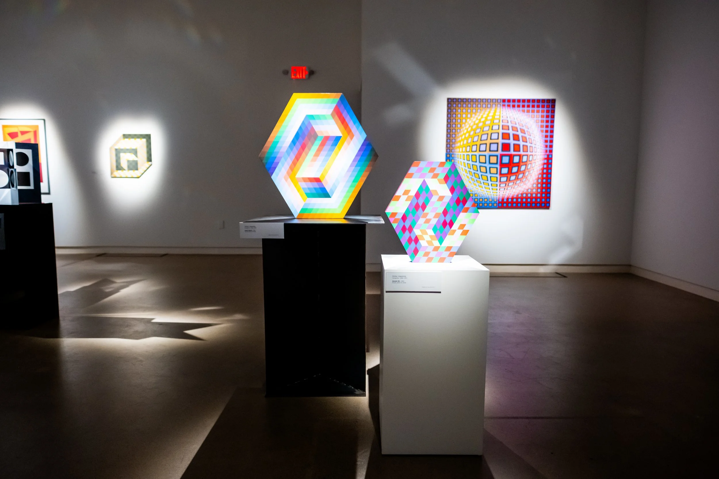 An art gallery display featuring colorful geometric light sculptures and paintings with geometric patterns on the wall, illuminated in a dimly lit room.