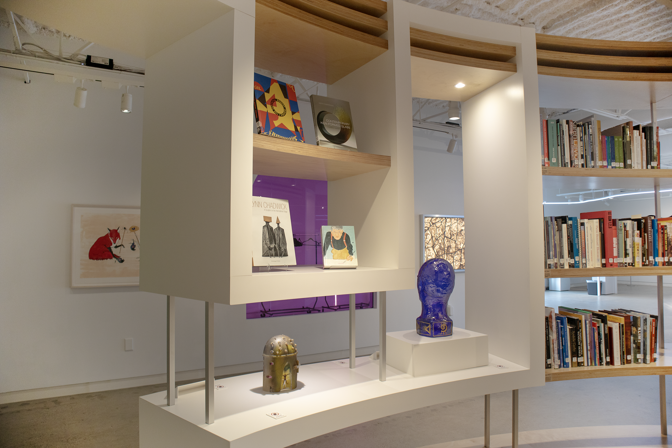 Interior of a modern library or gallery with a white shelving unit displaying books, artwork, and decorative objects including a blue vase and small sculpture, with additional bookshelves and a ceiling light in the background.