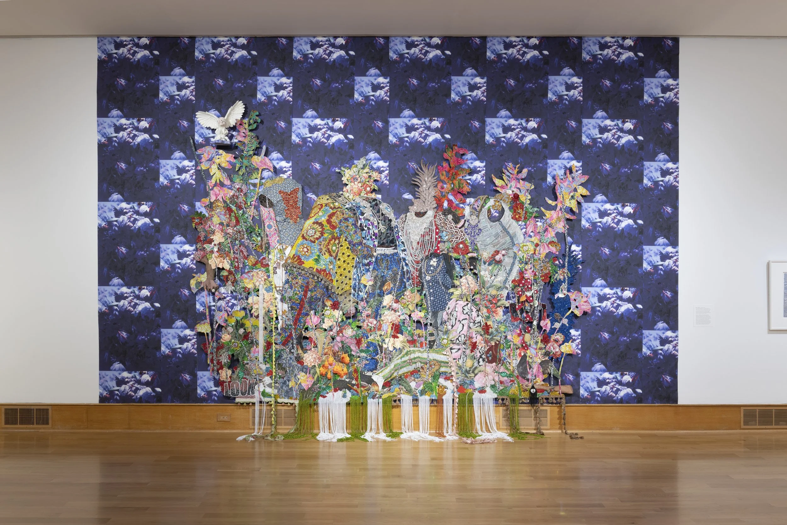 A mixed media art installation featuring embroidered and fabric artwork of elephants, set against a deep blue floral backdrop in a gallery space.