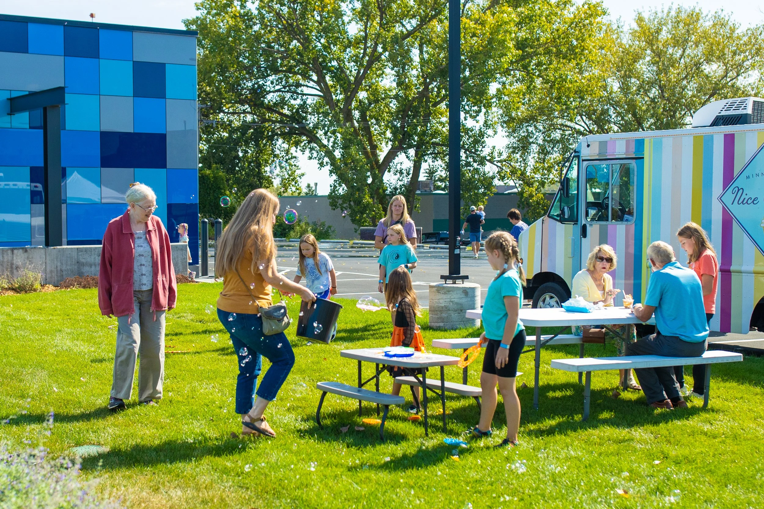 A group of people of varying ages blow bubbles on the CAT's front lawn, in the background some people eat ice cream.