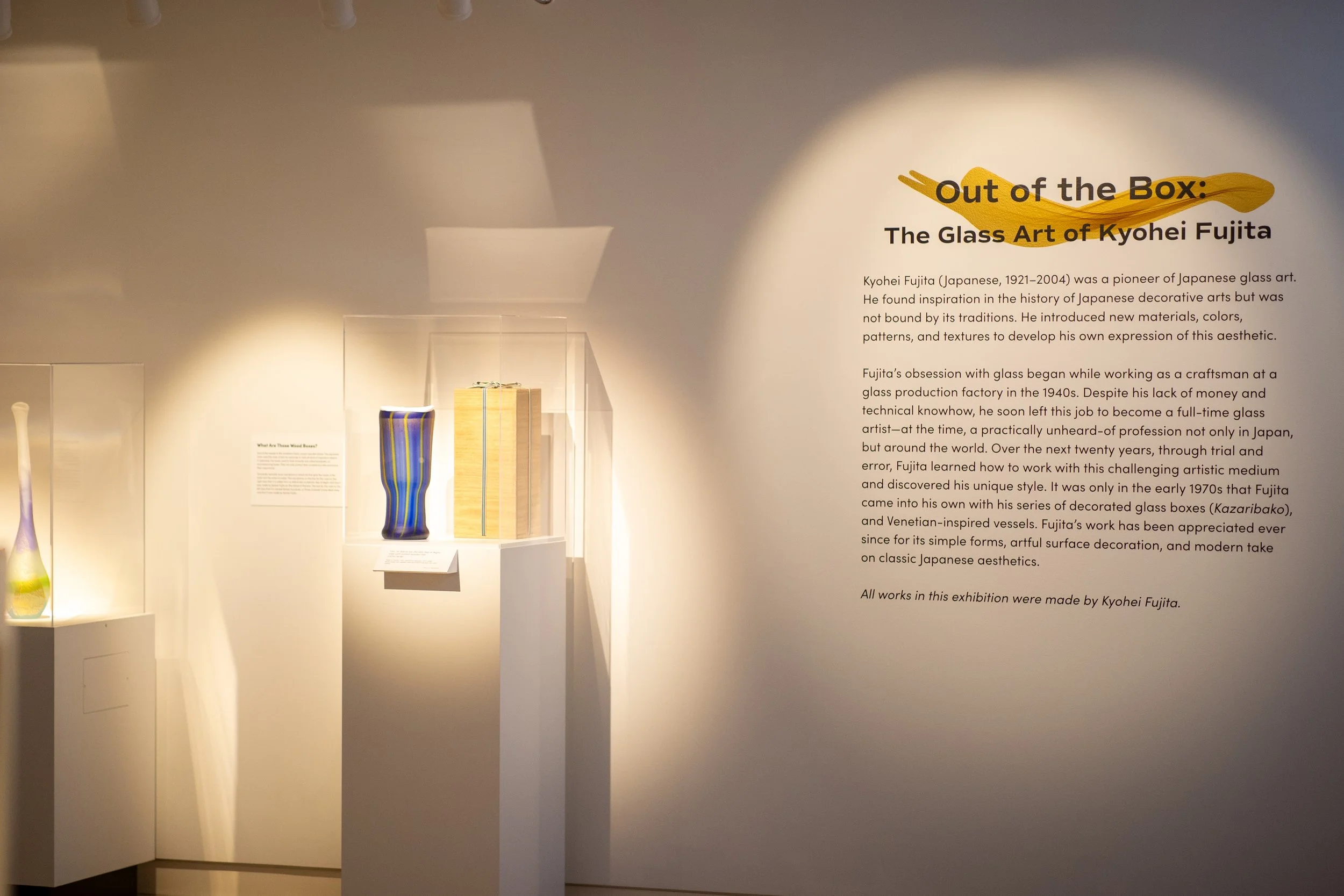 Exhibition display of glass art pieces including a tall blue and yellow striped vase and a wooden box, with informational plaques and descriptive text about Japanese glass artist Kyohei Fujita.