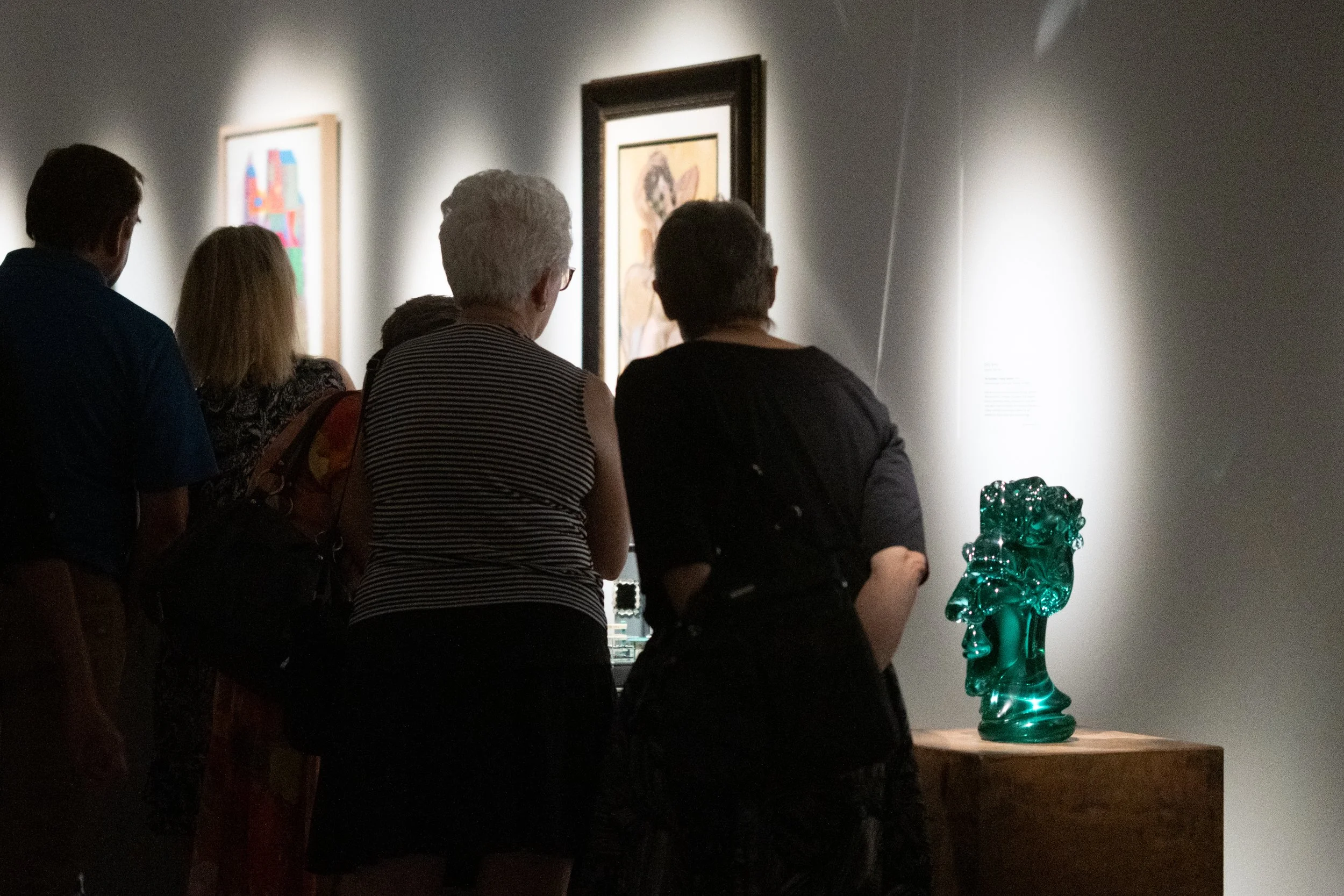 Group of people viewing artwork in a gallery, with a bright turquoise glass sculpture on display in the foreground.