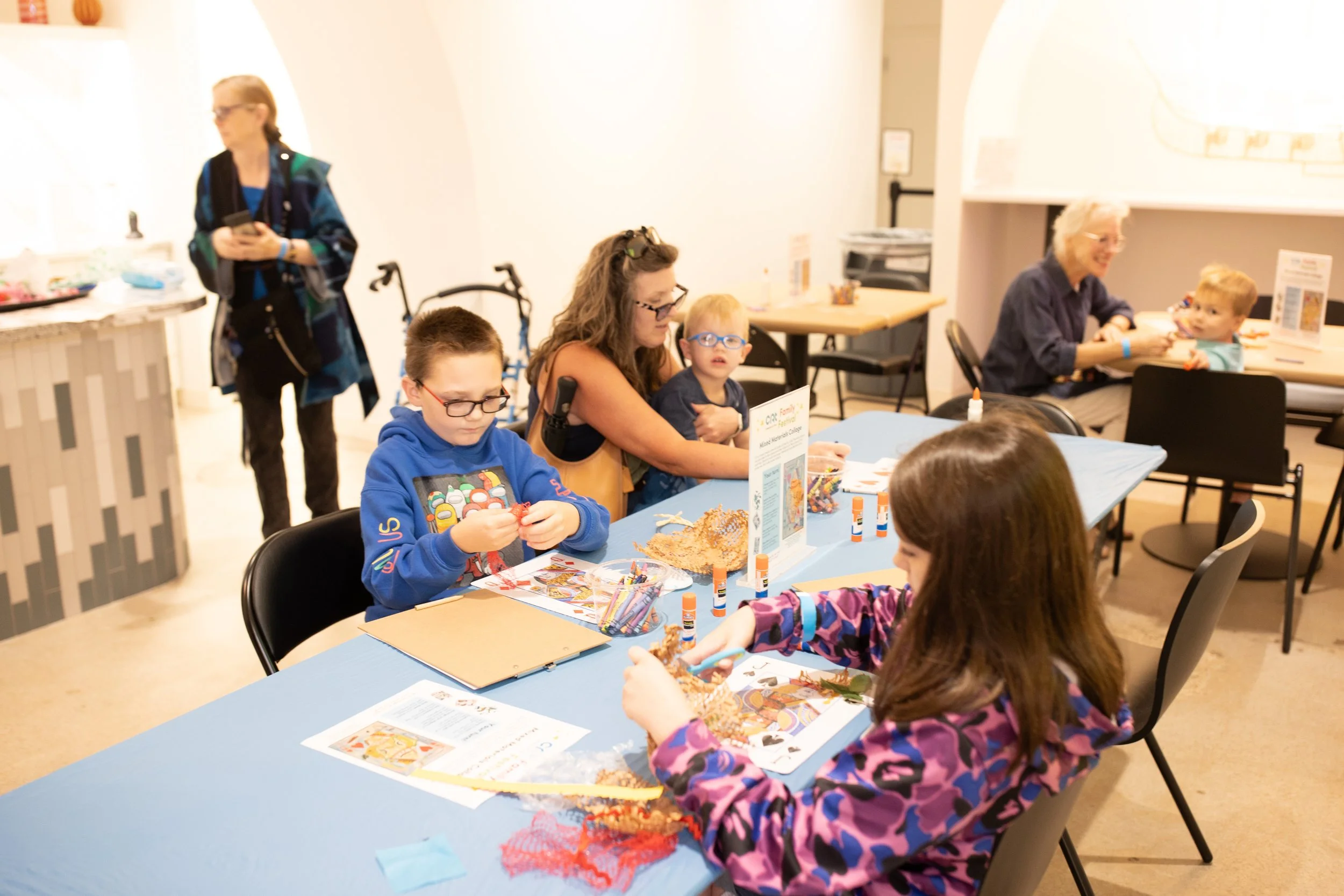 Adults and children gather around a table covered with various craft and art supplies.