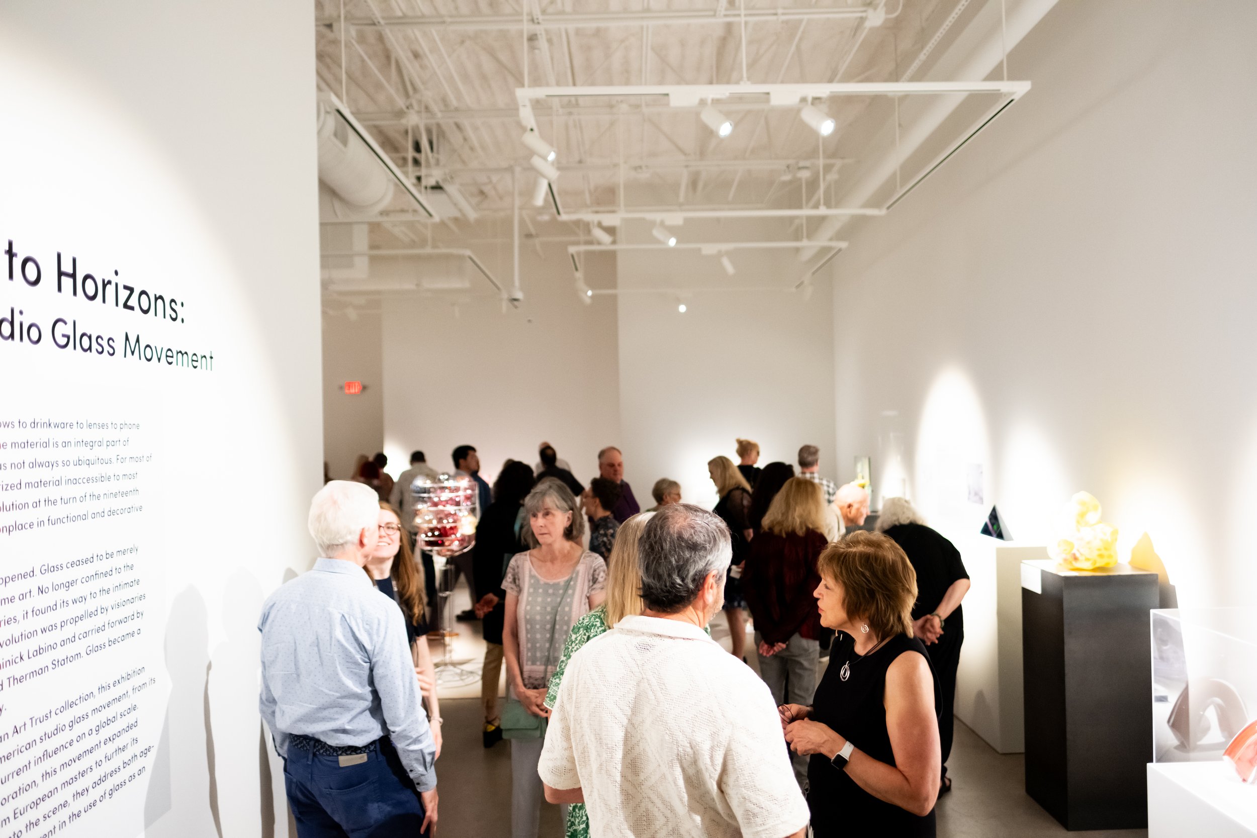 People gather in a gallery, there are glass sculptures on pedestals around them.