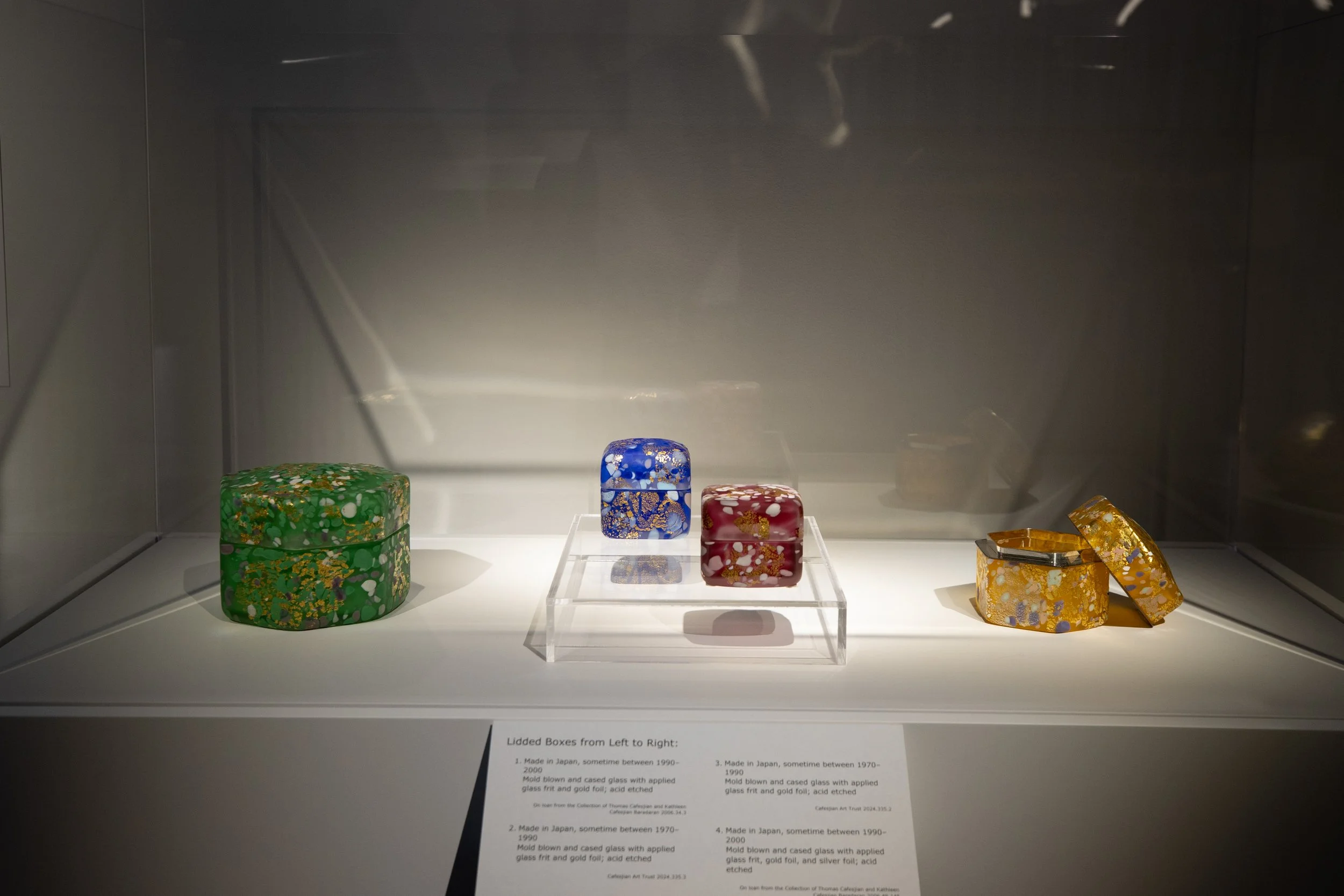 Display of four colorful, decorative, lacquered boxes arranged on a white shelf. The boxes are in green, blue, red, and yellow, each with gold and white patterns. A small sign below provides information about their origins and descriptions.