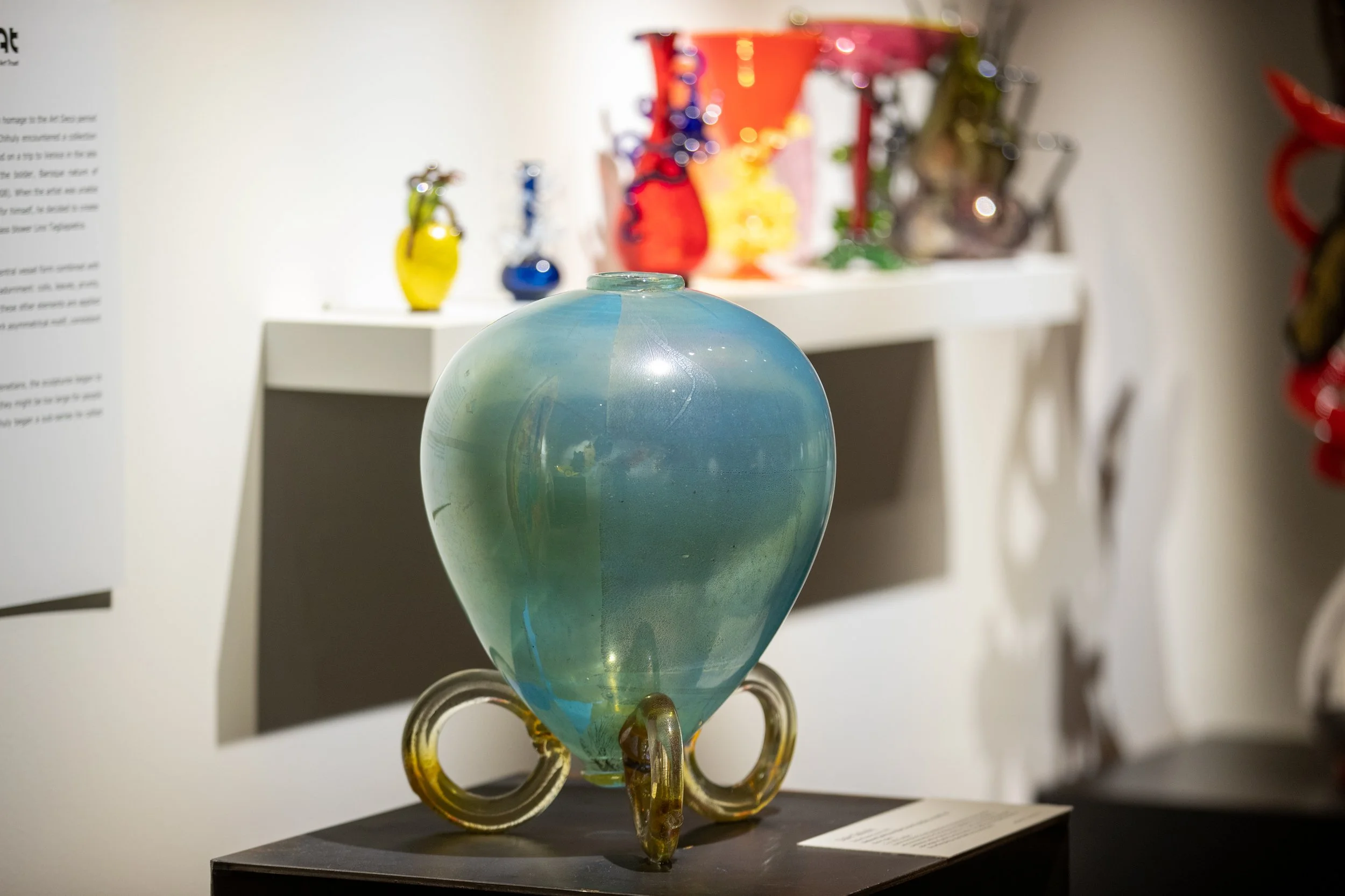 A glass art sculpture resembling a whimsical creature with a rounded, iridescent blue-green body, gold accents, and decorative rings at its base on display in a museum or gallery setting.