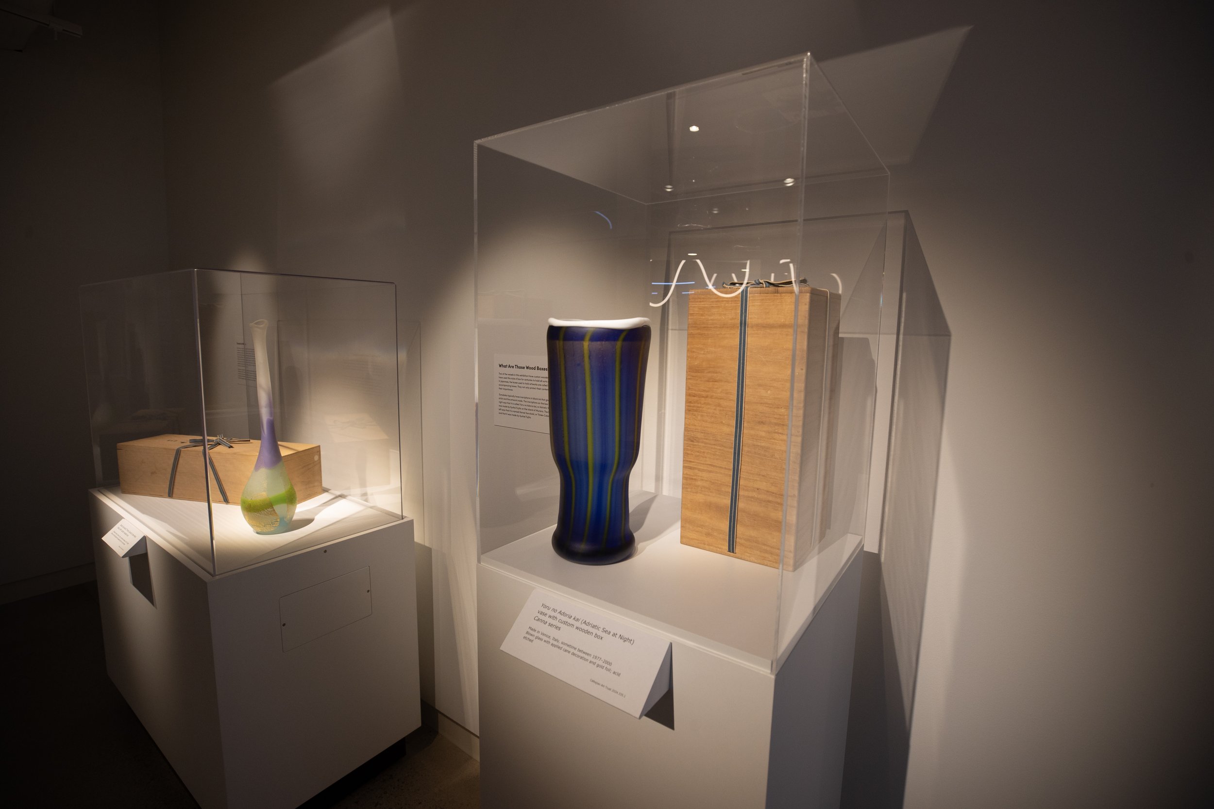 Display of colorful glass vases in a museum exhibit, with informational placards, enclosed in glass cases.