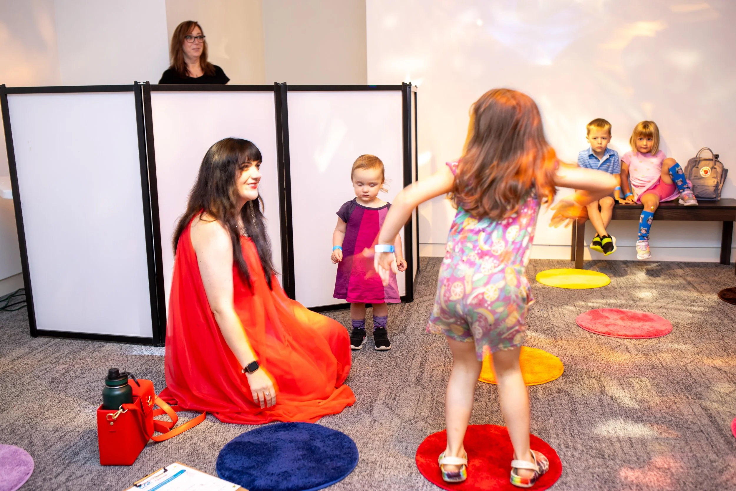 An adult kneels next to two children dancing; a DJ is in the background.