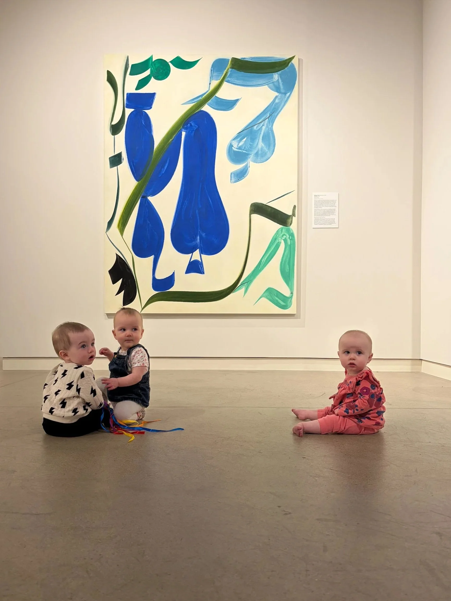 Three babies seated on the gallery floor in front of a large abstract painting.