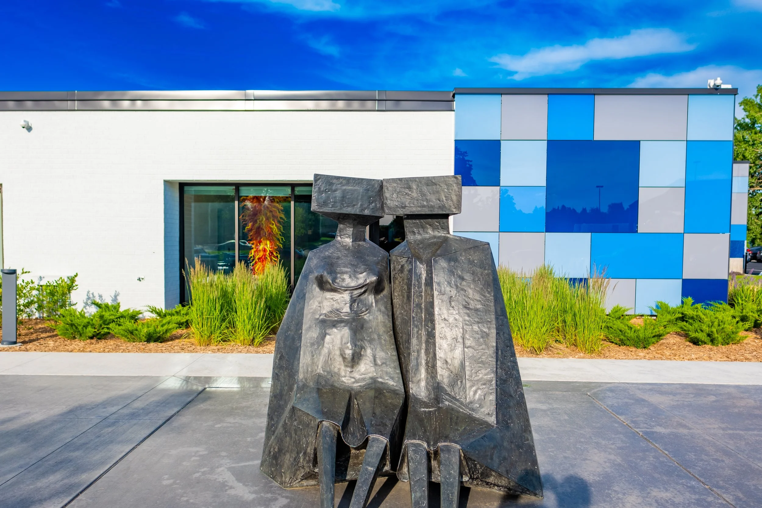 Art sculpture of two abstract human figures in front of a modern building with blue and gray panels, green shrubs, and a clear blue sky.