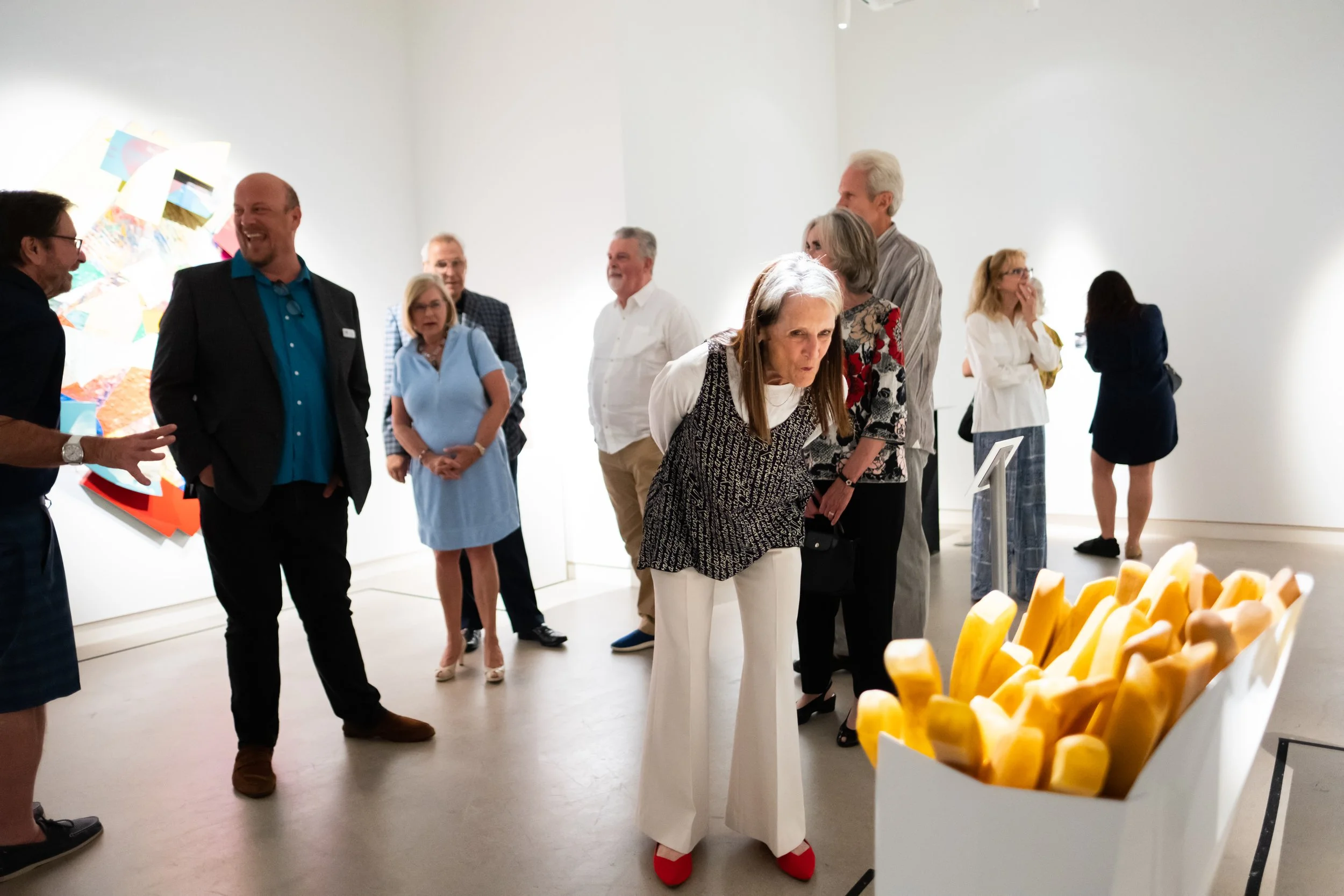 A group of people gathered in an art gallery, engaging in conversation. There are paintings on the walls and an installation of cheese in the foreground.