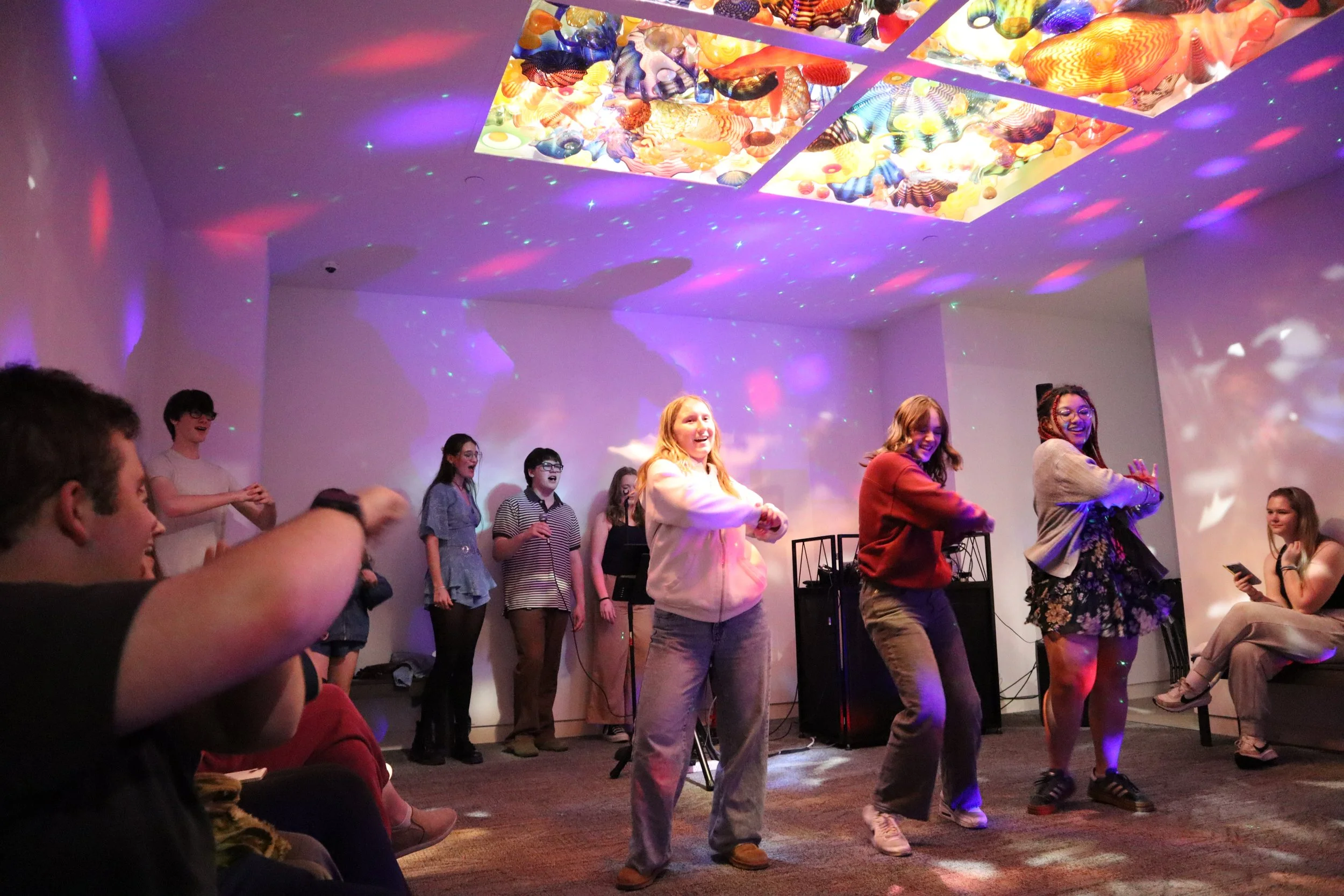 Three teenagers dancing under colorful lights while others watch and sing along.