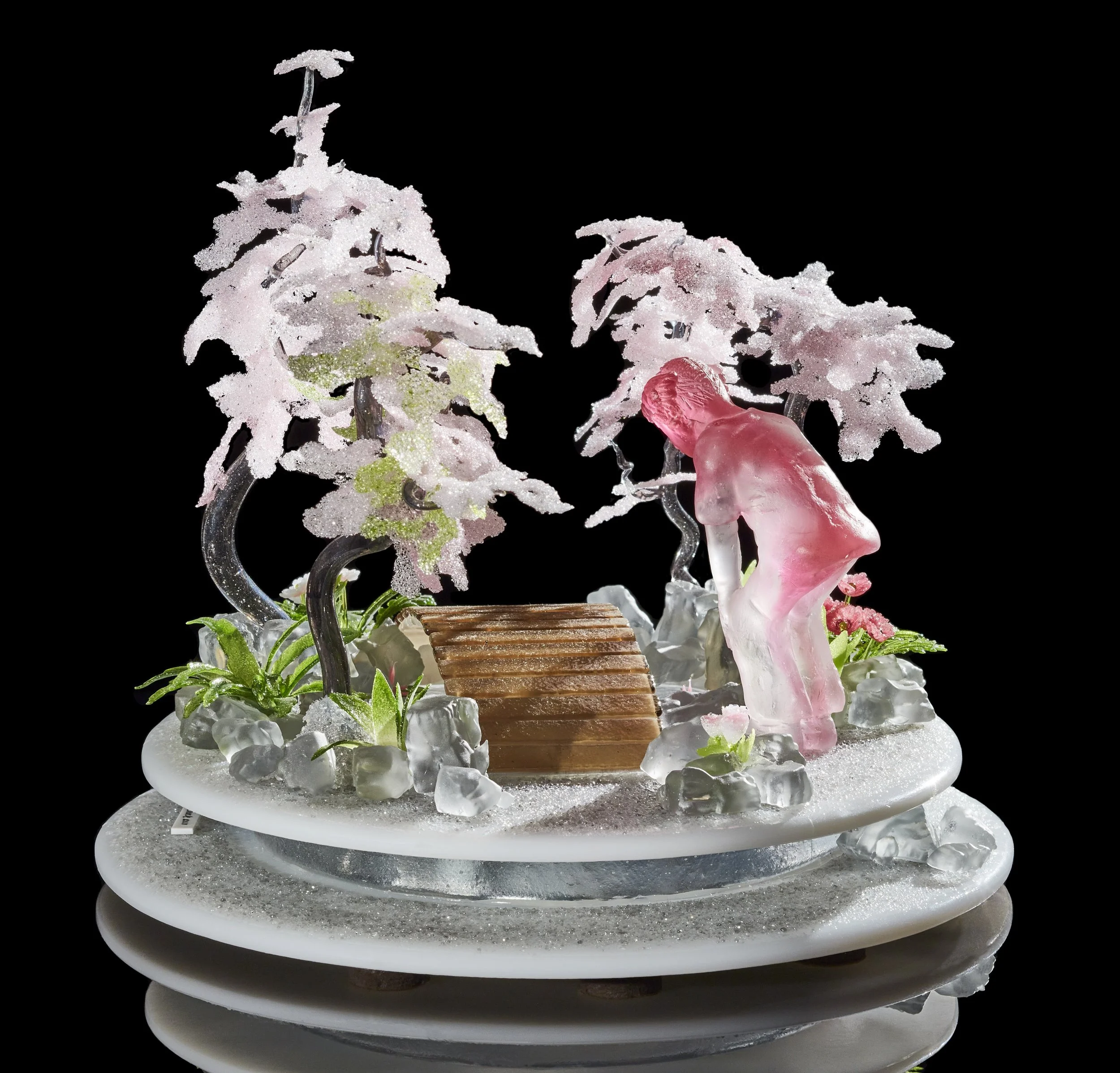 Glass sculpture on a round base of trees and a small bridge with a pink human figure bending to look at the water.