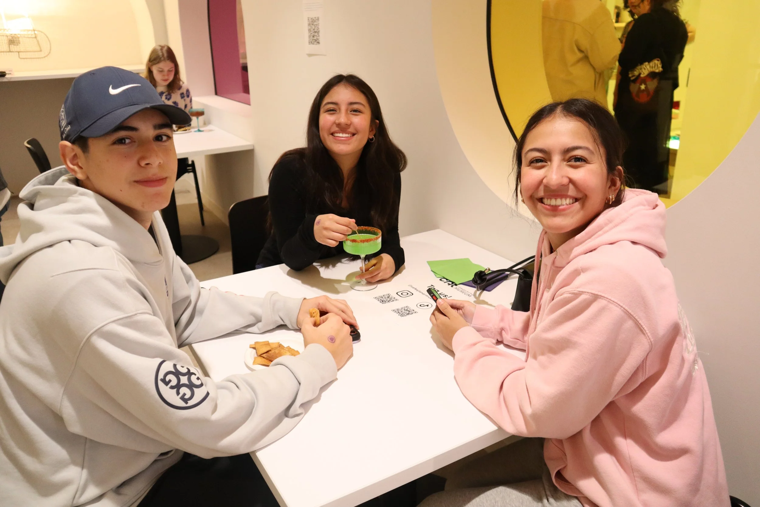 Three teenagers smiling for the camera while seated at a square table.
