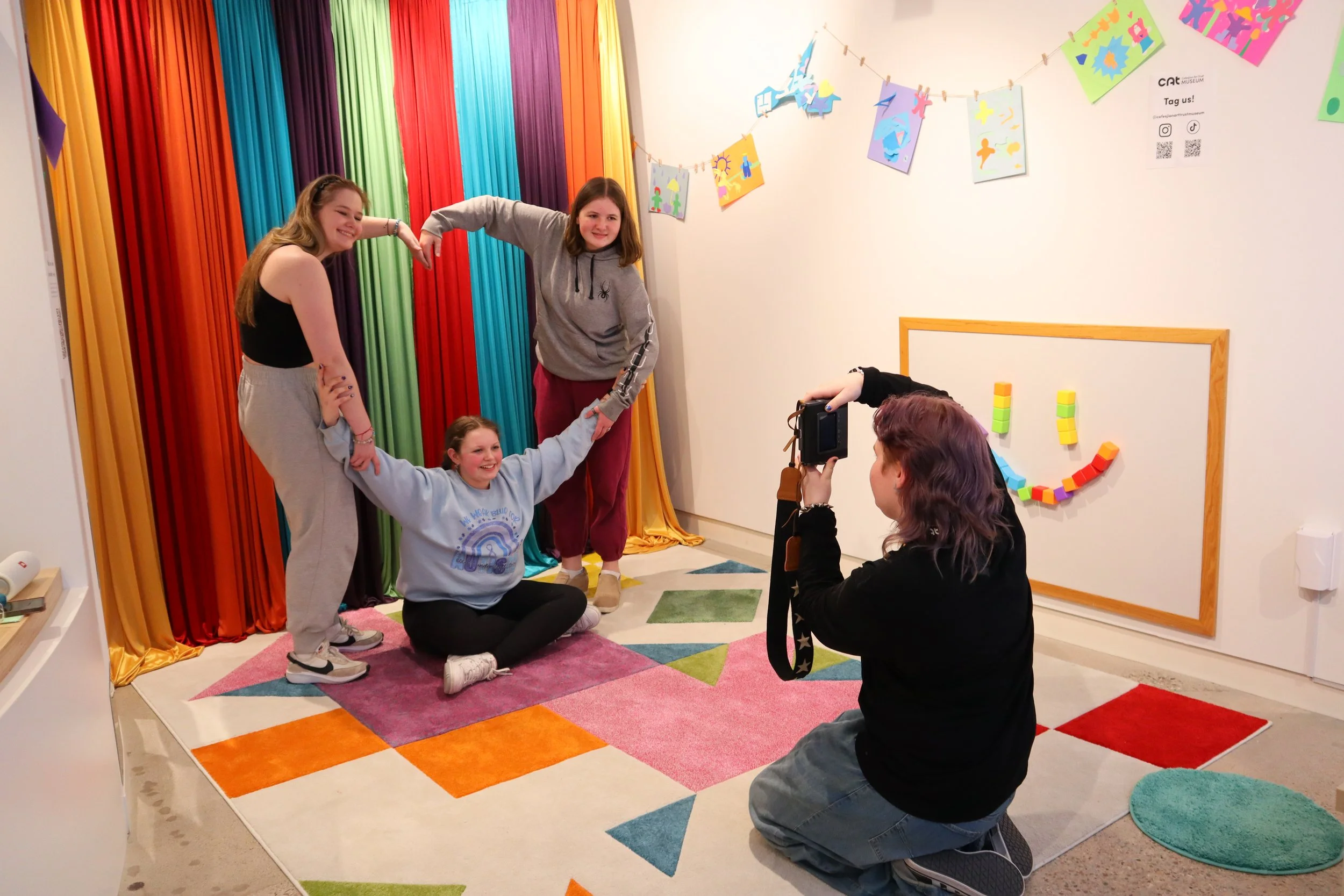 Three teenagers posing to make a heart shape with their arms while a fouth teeanger snaps their picture.