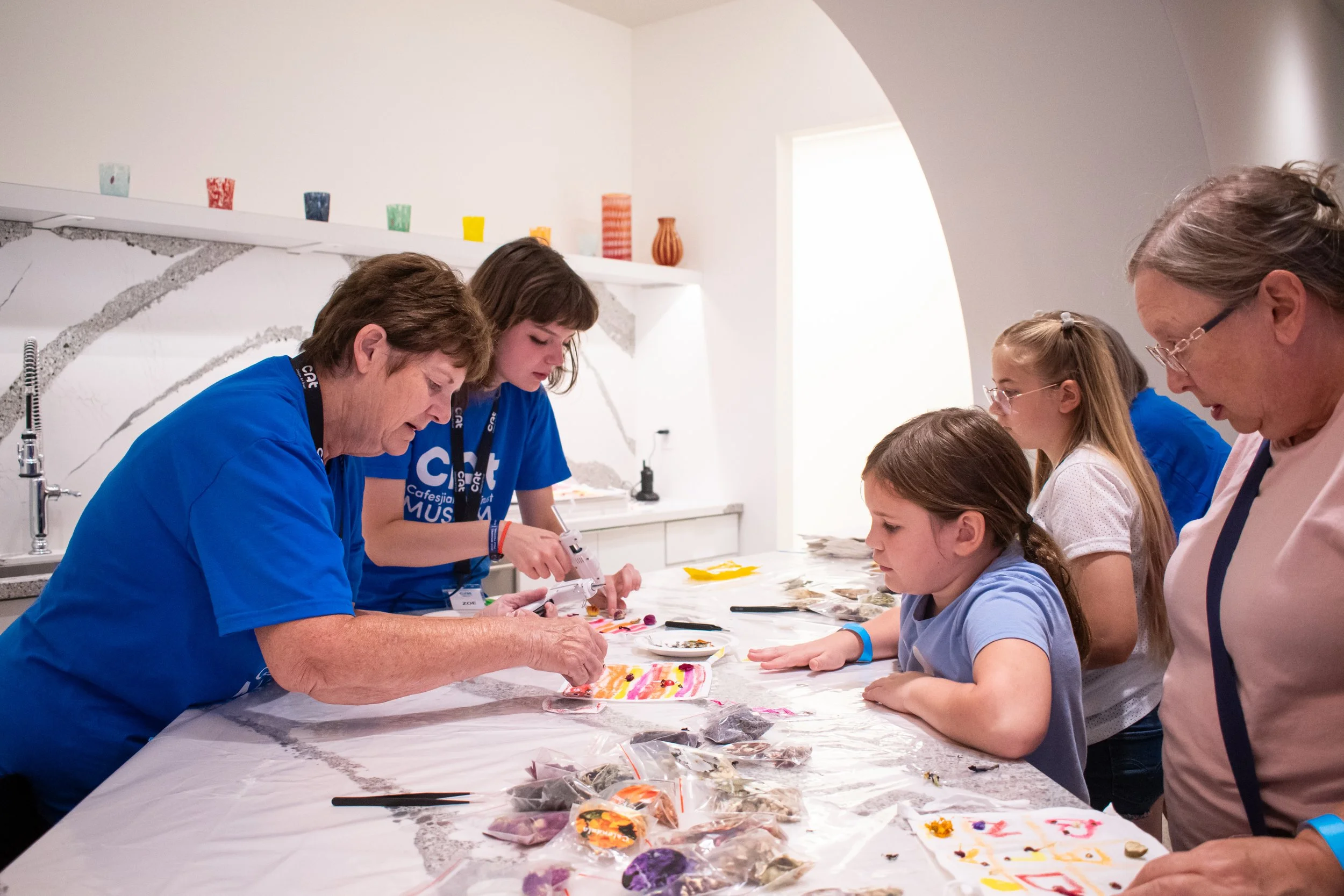 Two volunteers help visitors glue things to their art projects. 