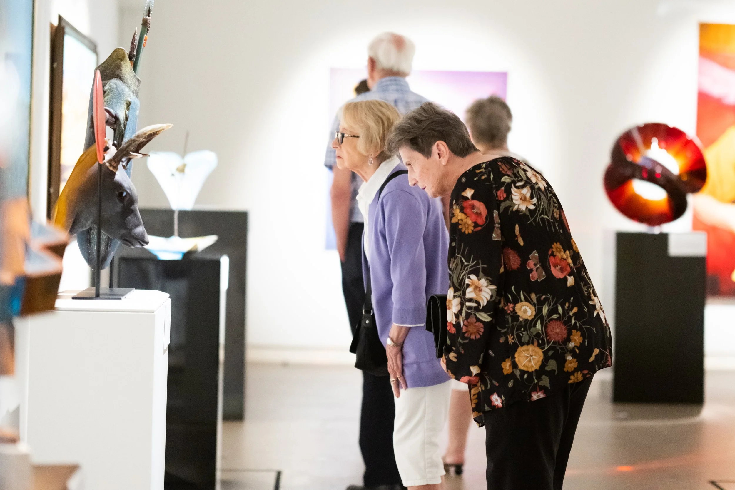 Several people are looking at art sculptures in a museum or gallery, standing side by side with a white wall in the background.