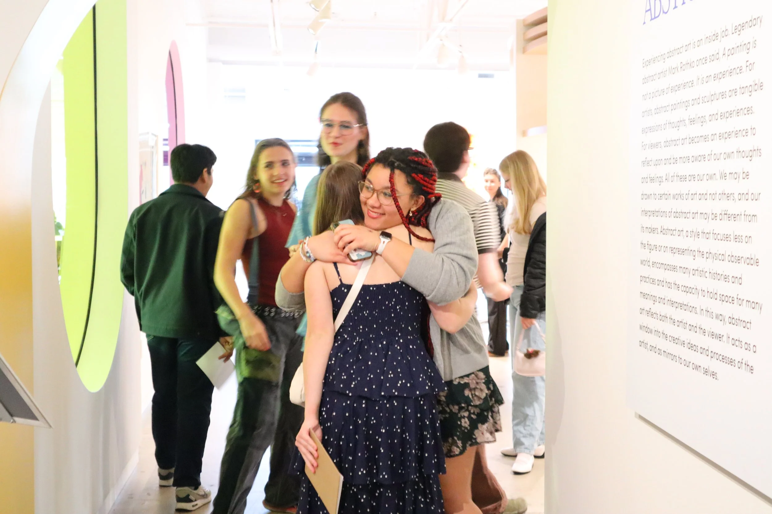 Two young adults hugging each other in greeting as others look on.