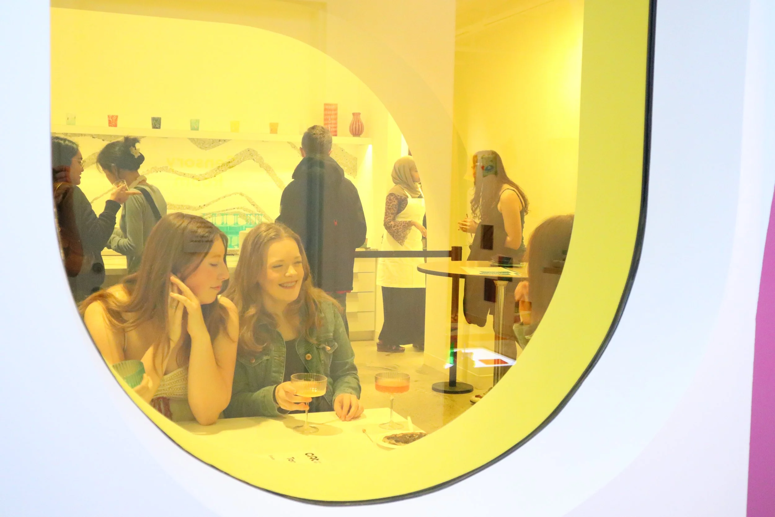 View through a bright yellow window of people gathered to eat snacks and drink mocktails.