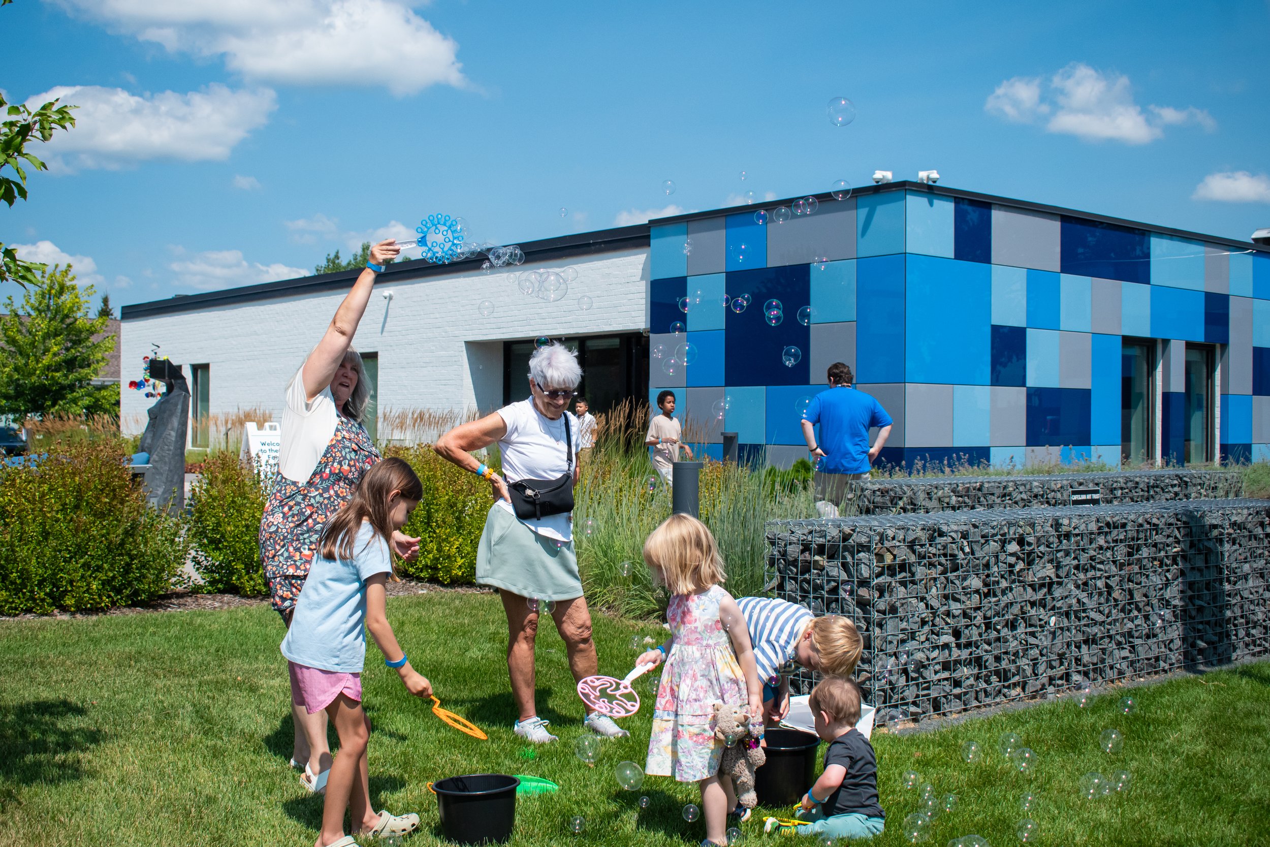 A group of children and adults gather on the lawn in front of the cat and blow bubbles. 