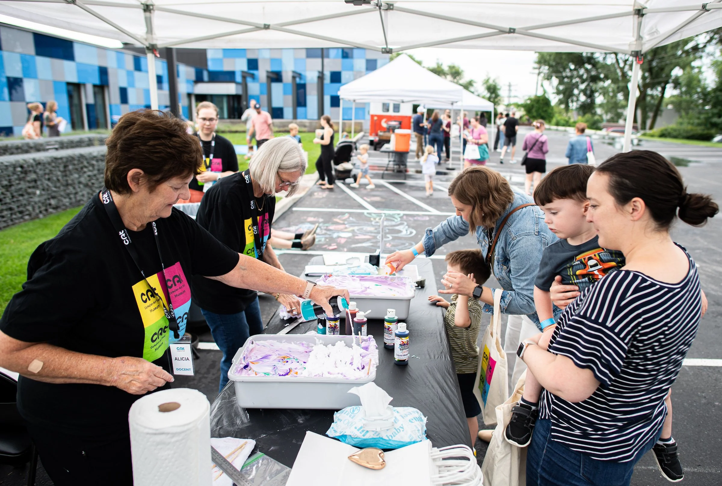 Families and volunteers working together on an art-making project outdoors under a white tent.