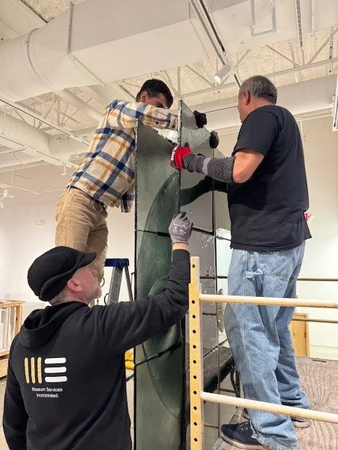 Three men work together to assemble the large abstract glass sculpture made of green-blue glass squares set into a metal grid.