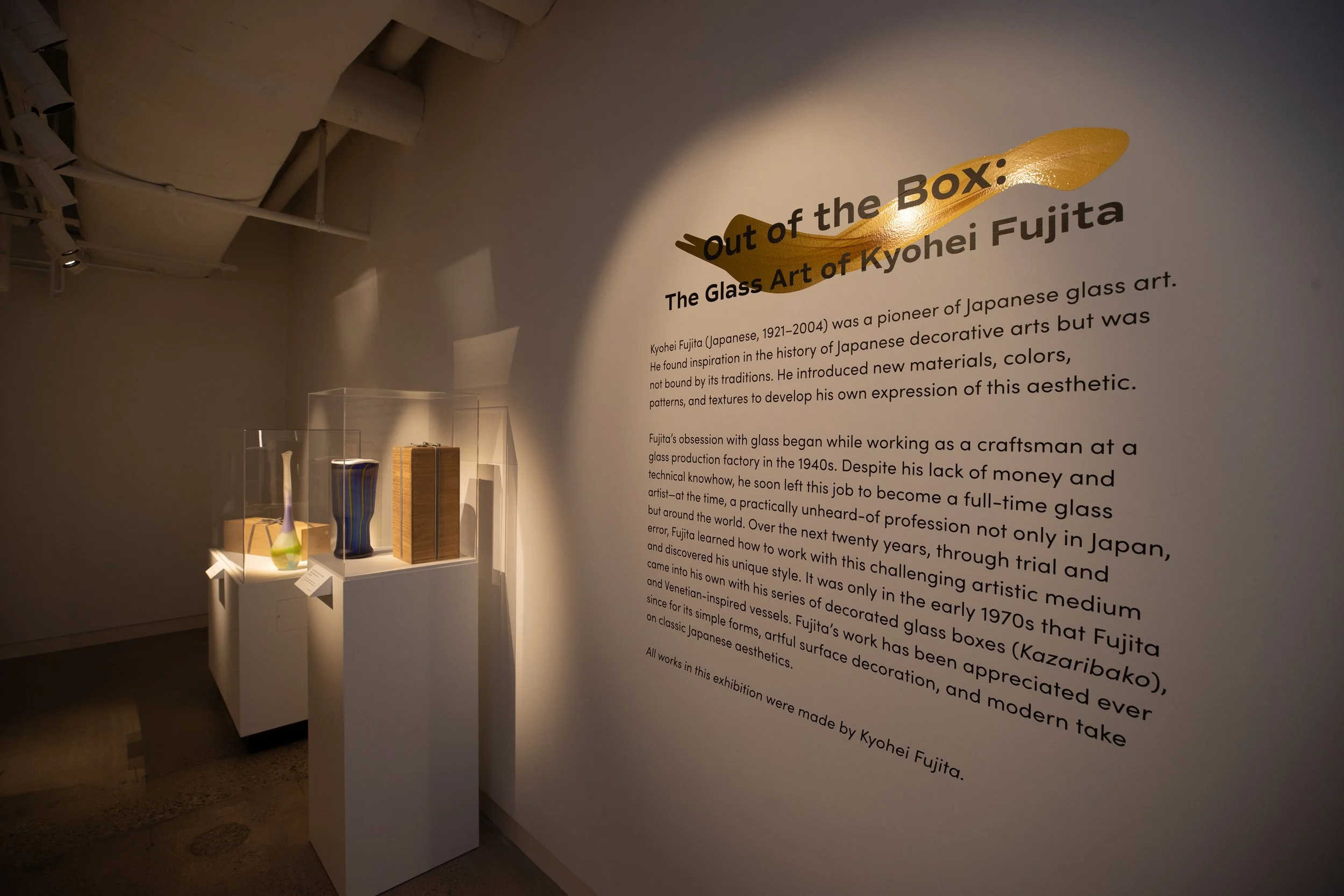 Exhibit wall with text about Kyohei Fujita and glass art, featuring a display case with colorful glass vases or sculptures.