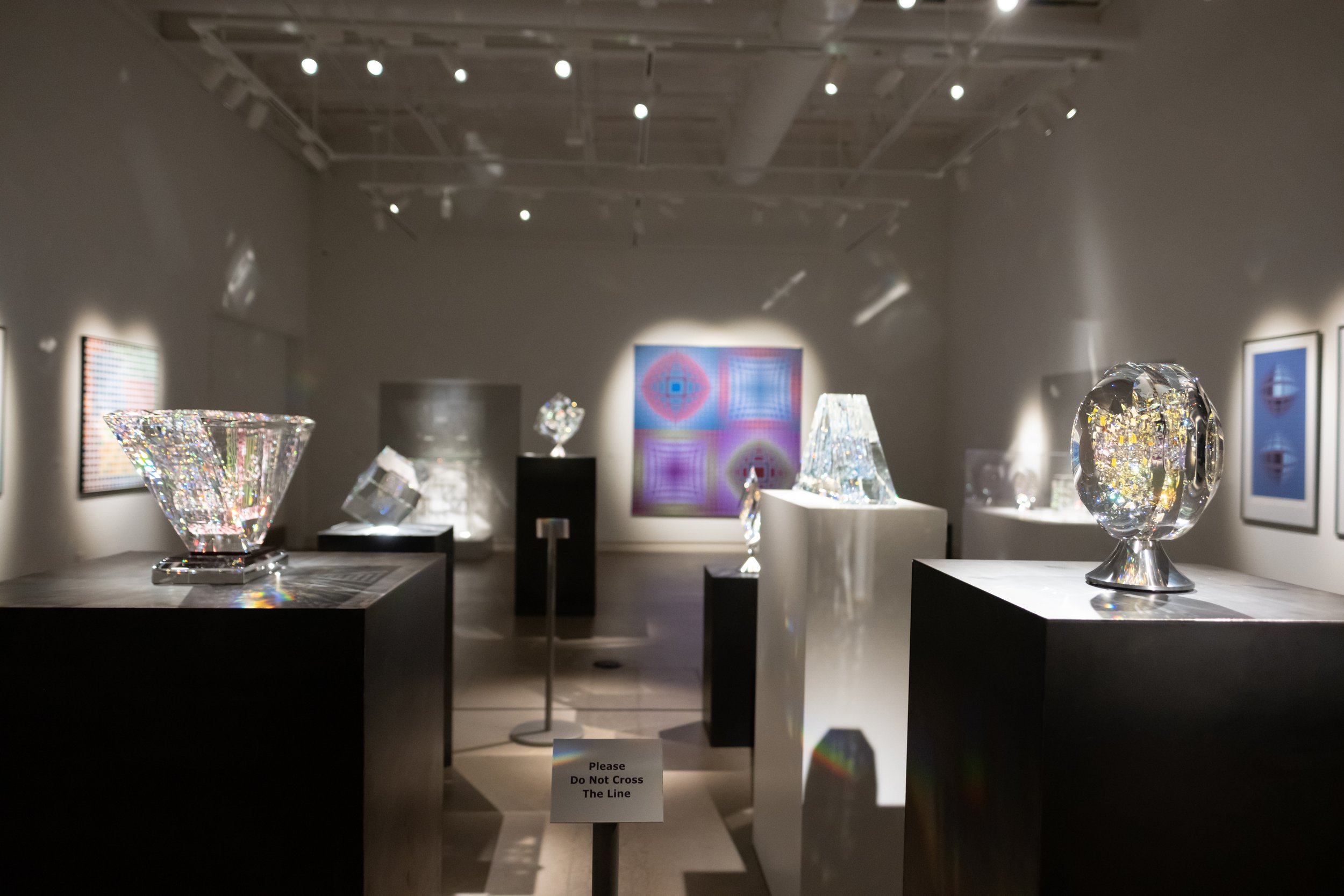 An exhibit of glass art sculptures displayed on pedestals in a gallery with framed artwork in the background. A sign in the foreground reads, 'Please Do Not Cross The Line.'
