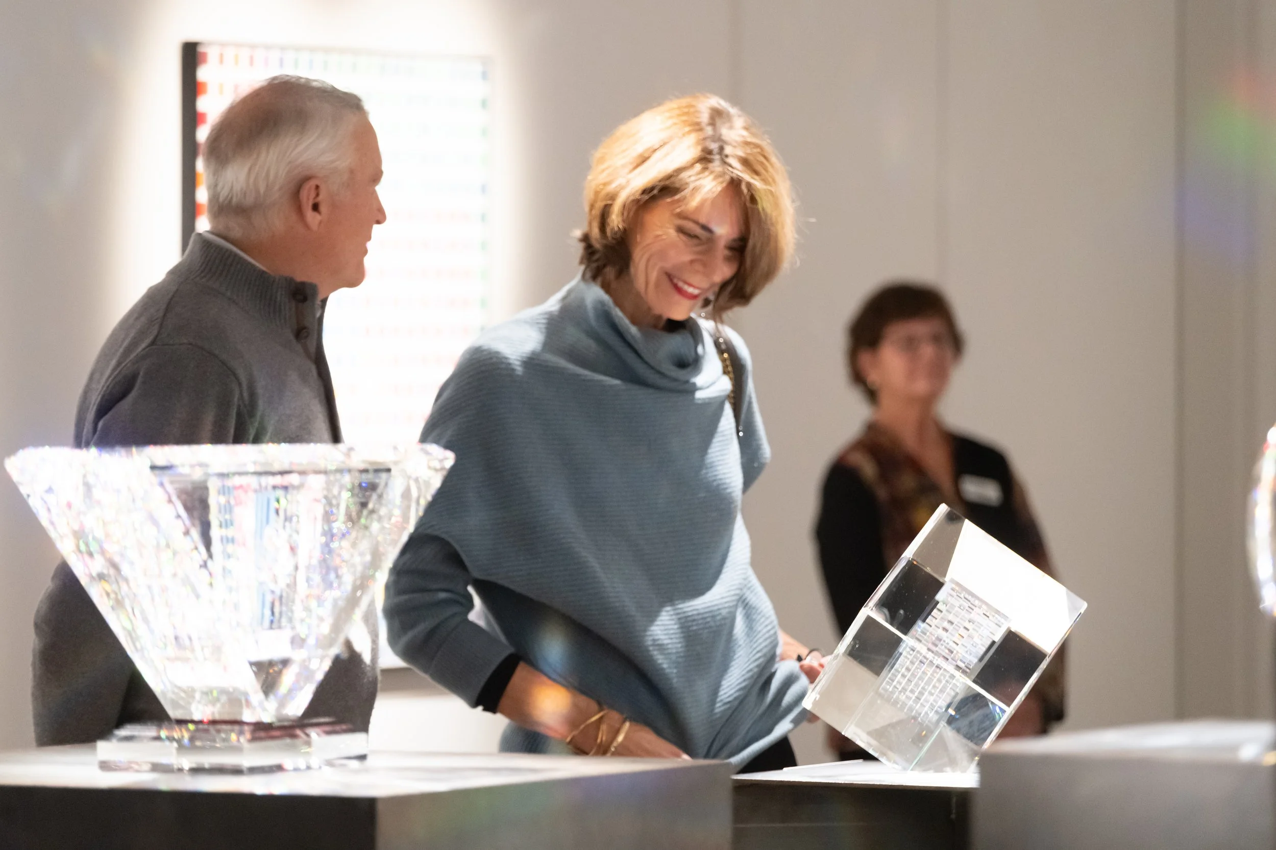 Woman with light brown hair smiling and looking at a display, standing next to a man with grey hair, both at an exhibition or event.
