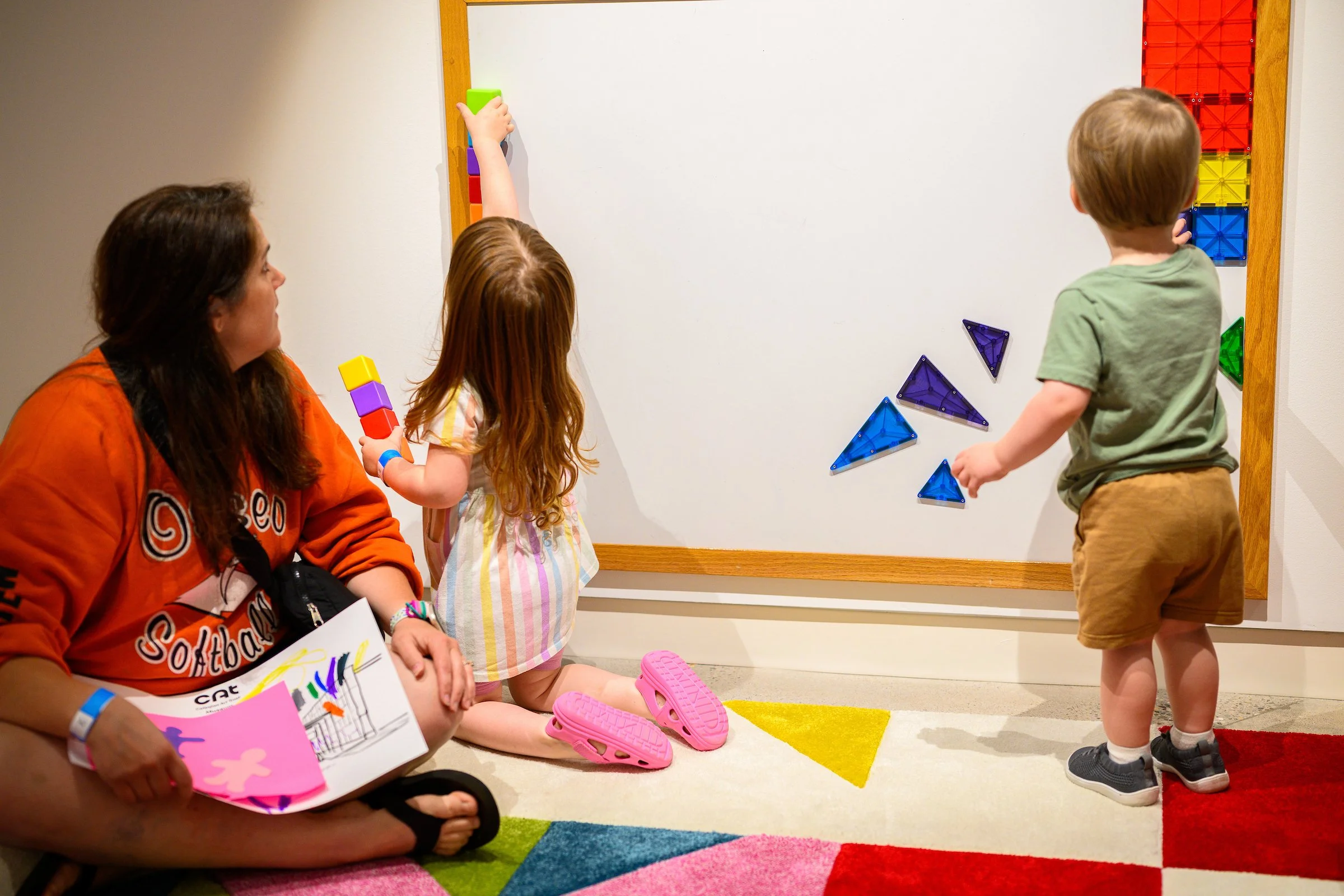 A mom looks on as two toddlers play with colorful magnets on a whiteboard.
