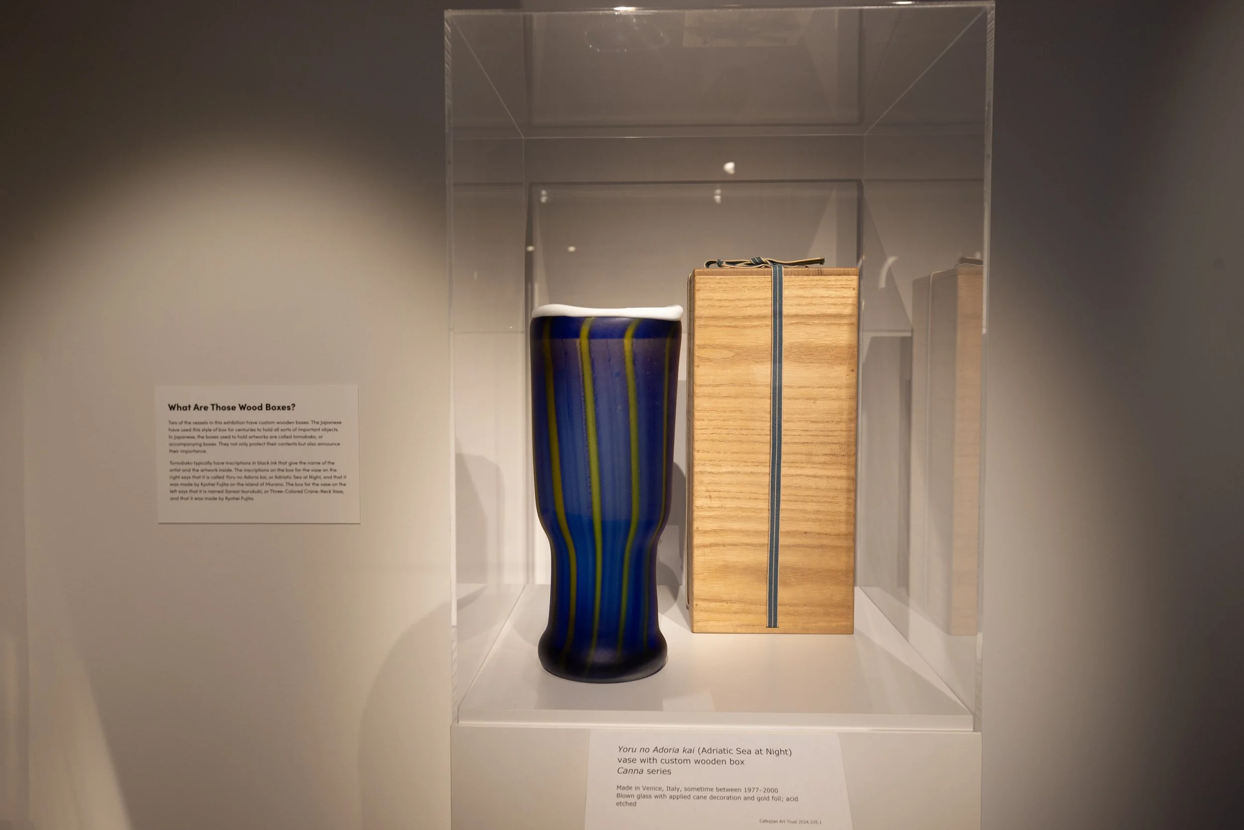 A tall, slender ceramic vase with a blue base color and wavy yellow and green vertical lines. The vase is displayed inside a glass case and is part of a museum exhibit. A label nearby provides information about the vase, indicating it is a Venetian b