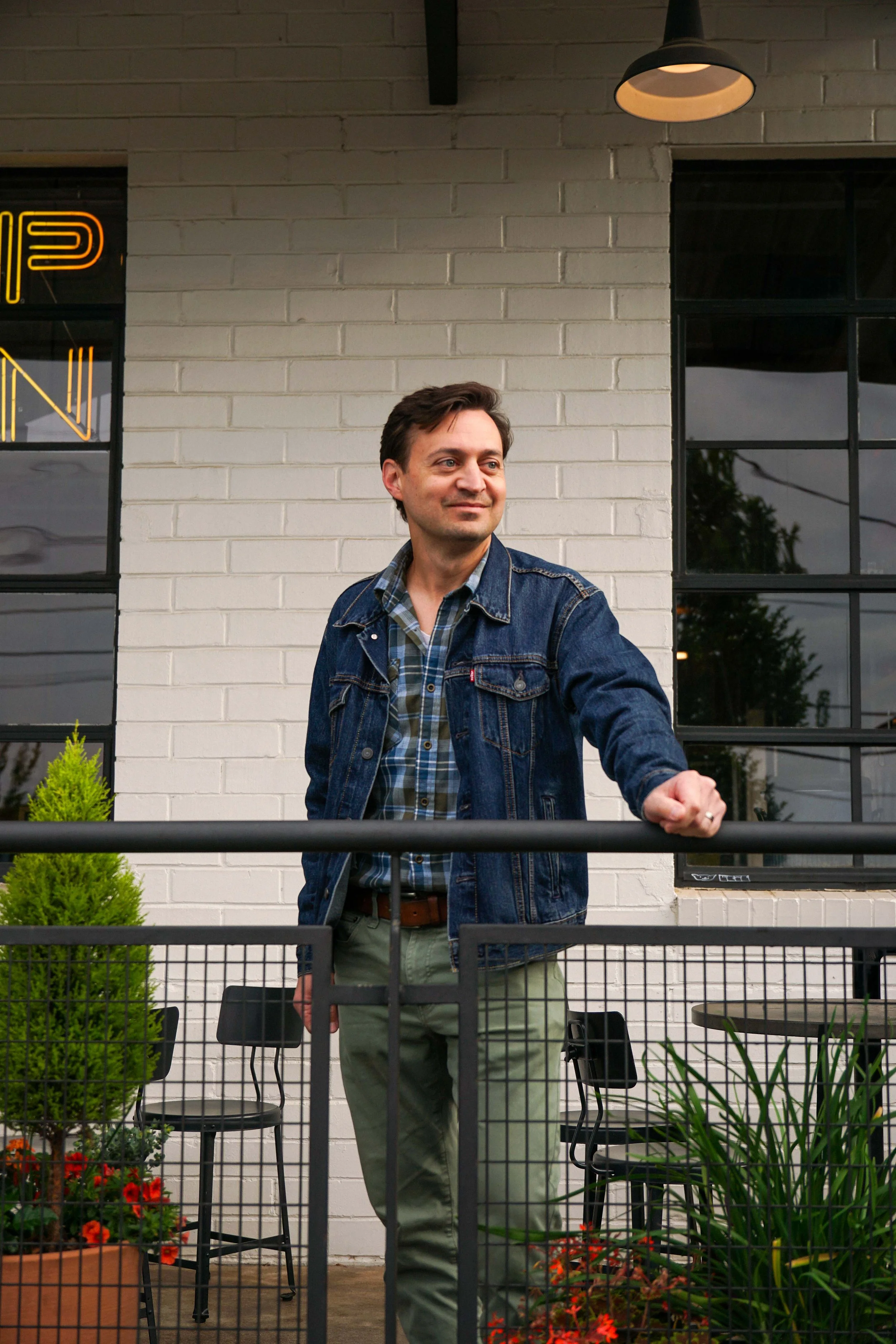 A man standing outdoors behind a metal railing, smiling and extending his fist, wearing a denim jacket and plaid shirt. There are chairs and plants in front of him, with a white brick wall and black-framed windows in the background.