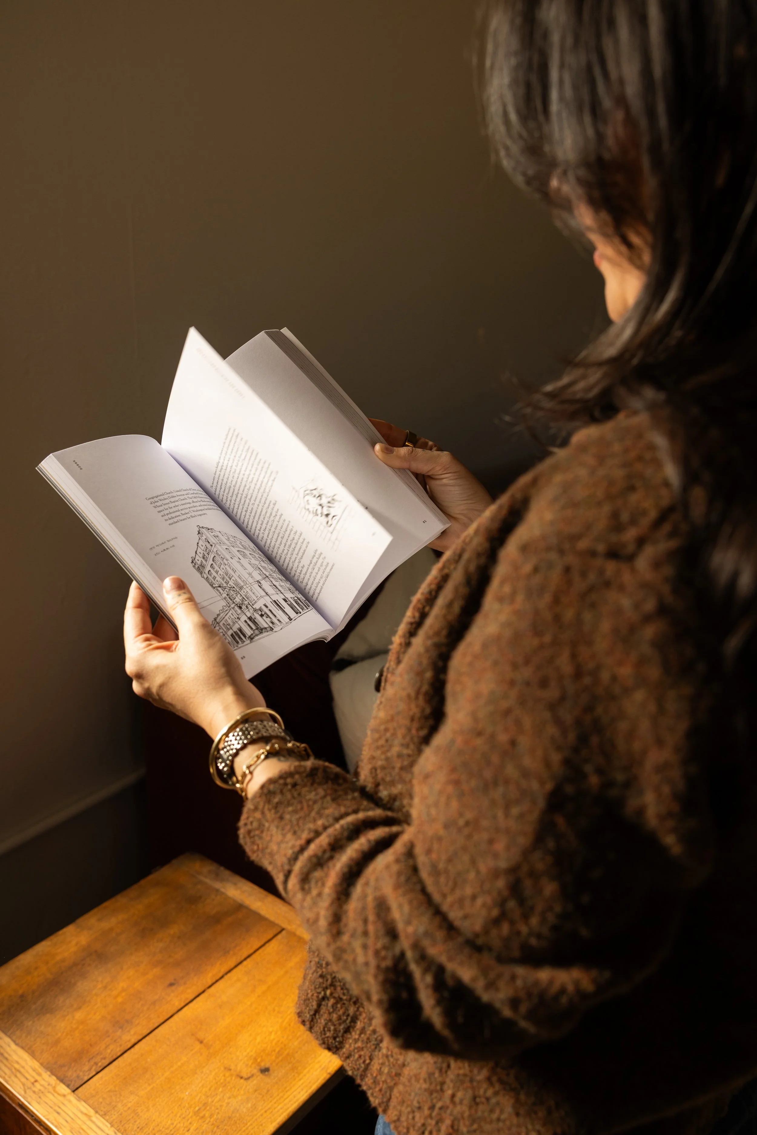 A woman with dark hair and jewelry, wearing a brown fuzzy sweater, is reading a book with a black and white illustration on one page. She is sitting at a wooden table.
