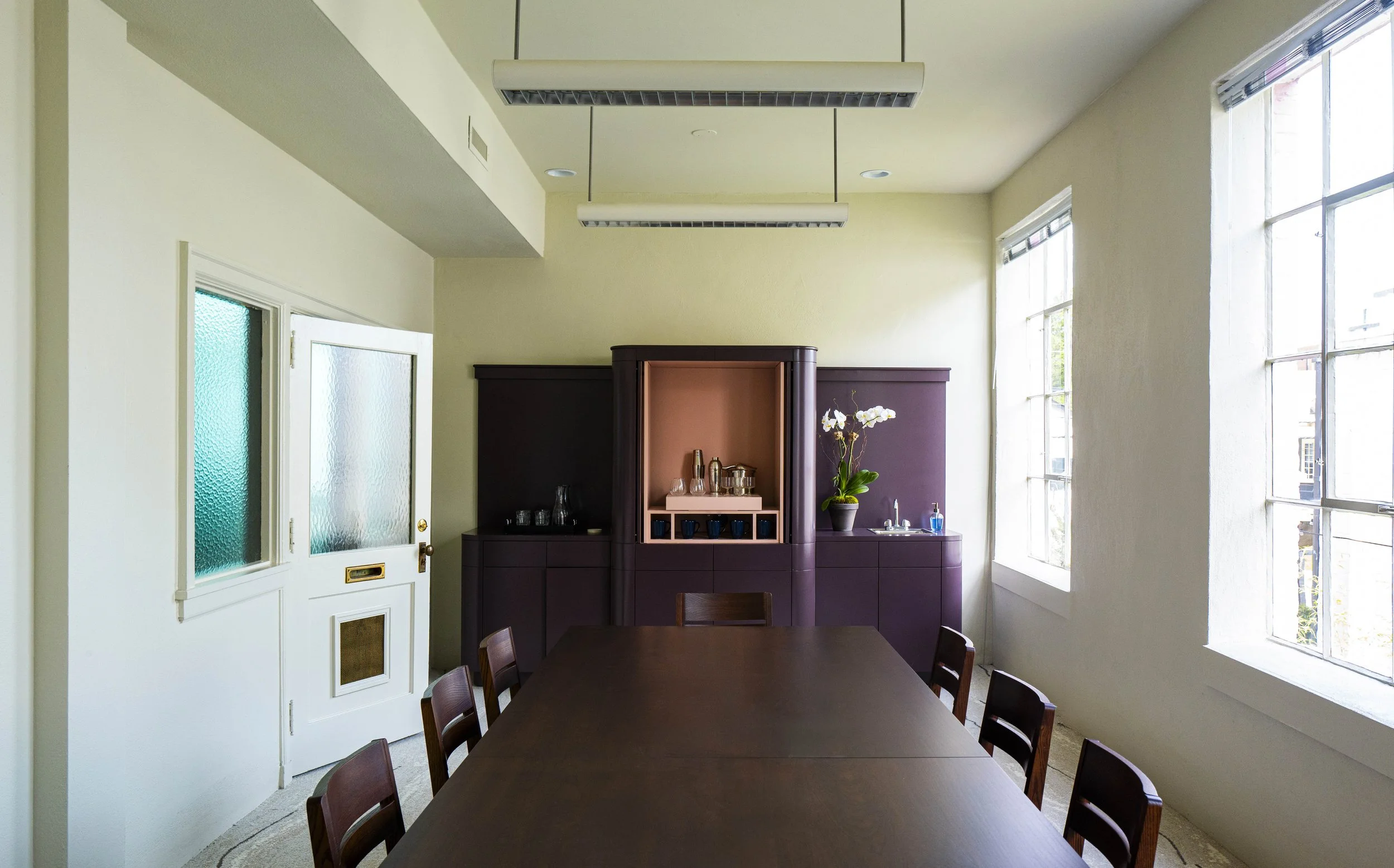 Interior of a dining room with a dark wood table and chairs, a purple kitchen cabinet with glassware and a potted orchid, large windows allowing natural light, and a white door with frosted glass panels.