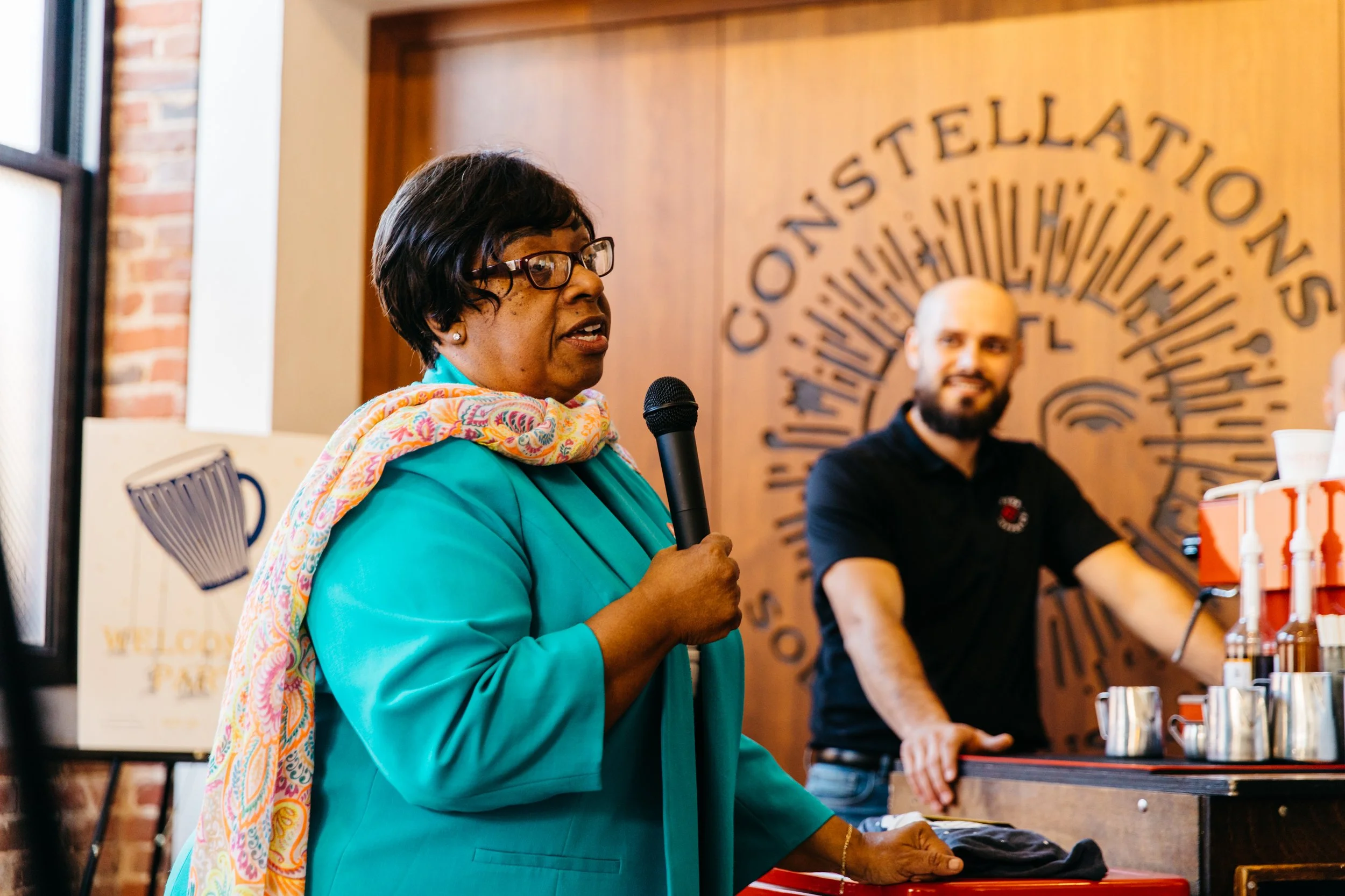 An African American woman speaking into a microphone at an indoor event, with a man sitting behind her at a table. The background features a wooden wall with the words 'Stellato' and other text curving around a circular design.