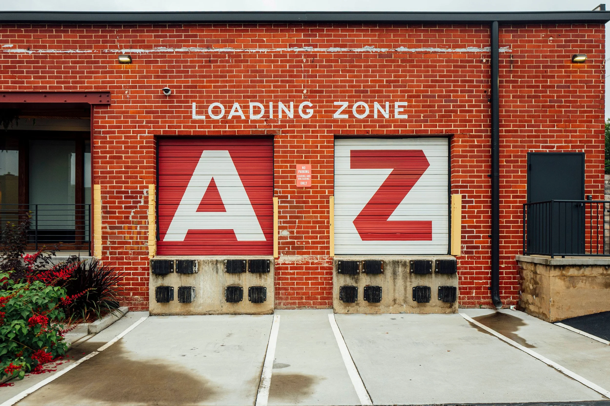 A red brick building with a sign reading 'Loading Zone' above two garage doors labeled with large red and white letters 'A' and 'Z'. Parking spaces in front marked with white lines and a small garden with red flowers on the left.