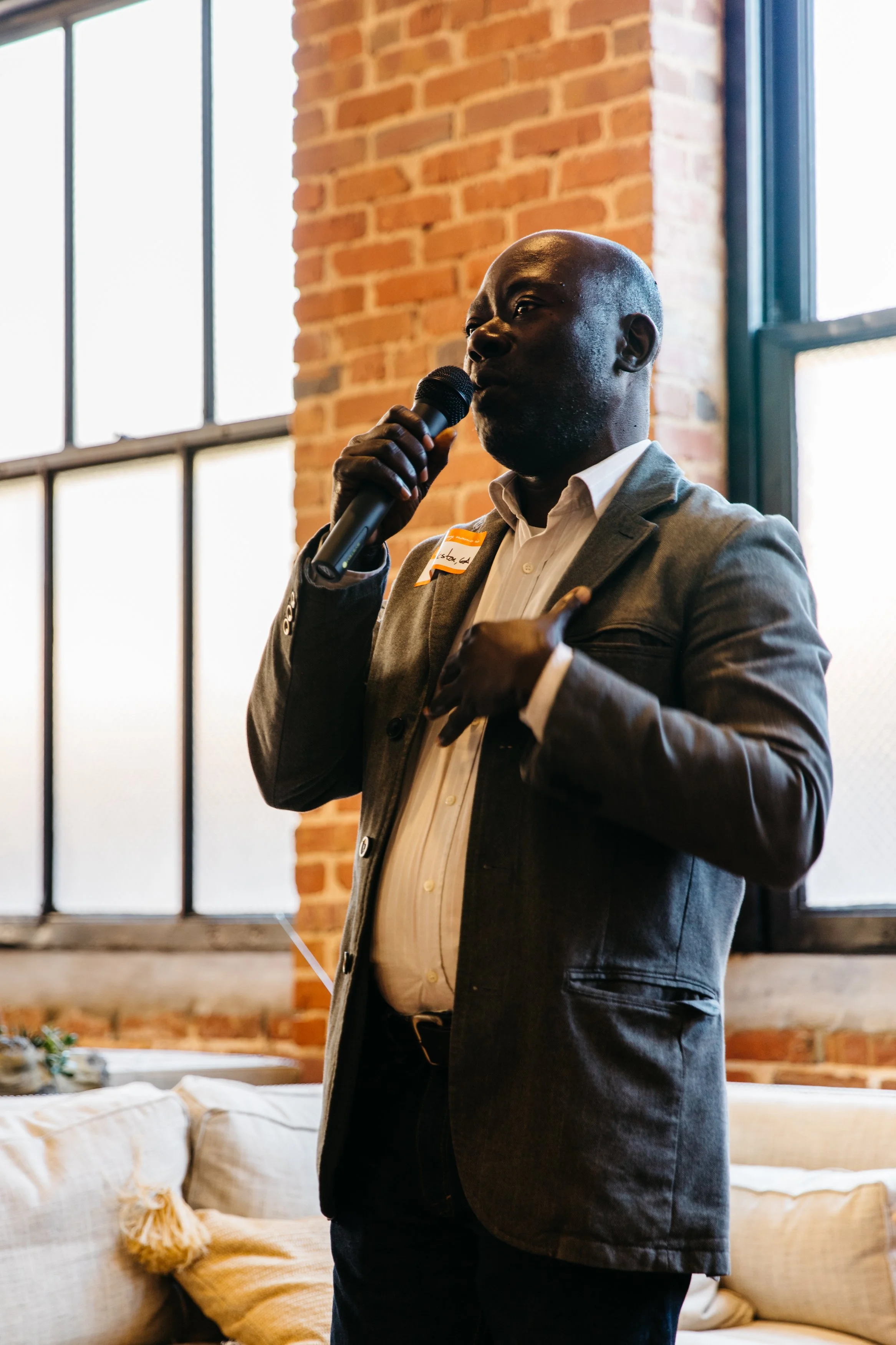 Man in gray blazer speaking into a microphone inside a room with exposed brick wall and large windows.