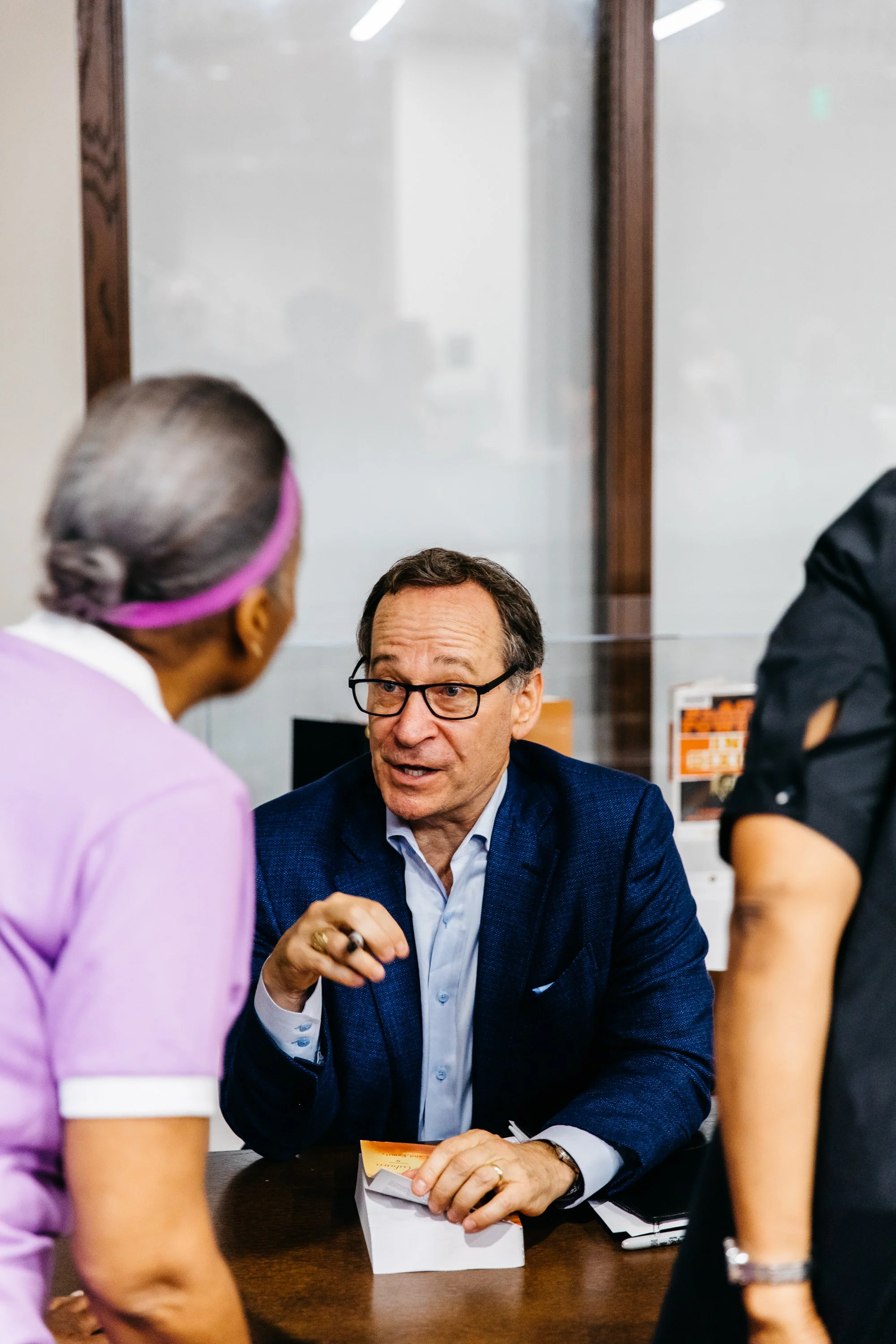 A man in a blue blazer and glasses is seated at a table, engaged in conversation with three women, one in light purple scrubs and two in dark clothing, in an indoor setting that appears to be an office or conference room.