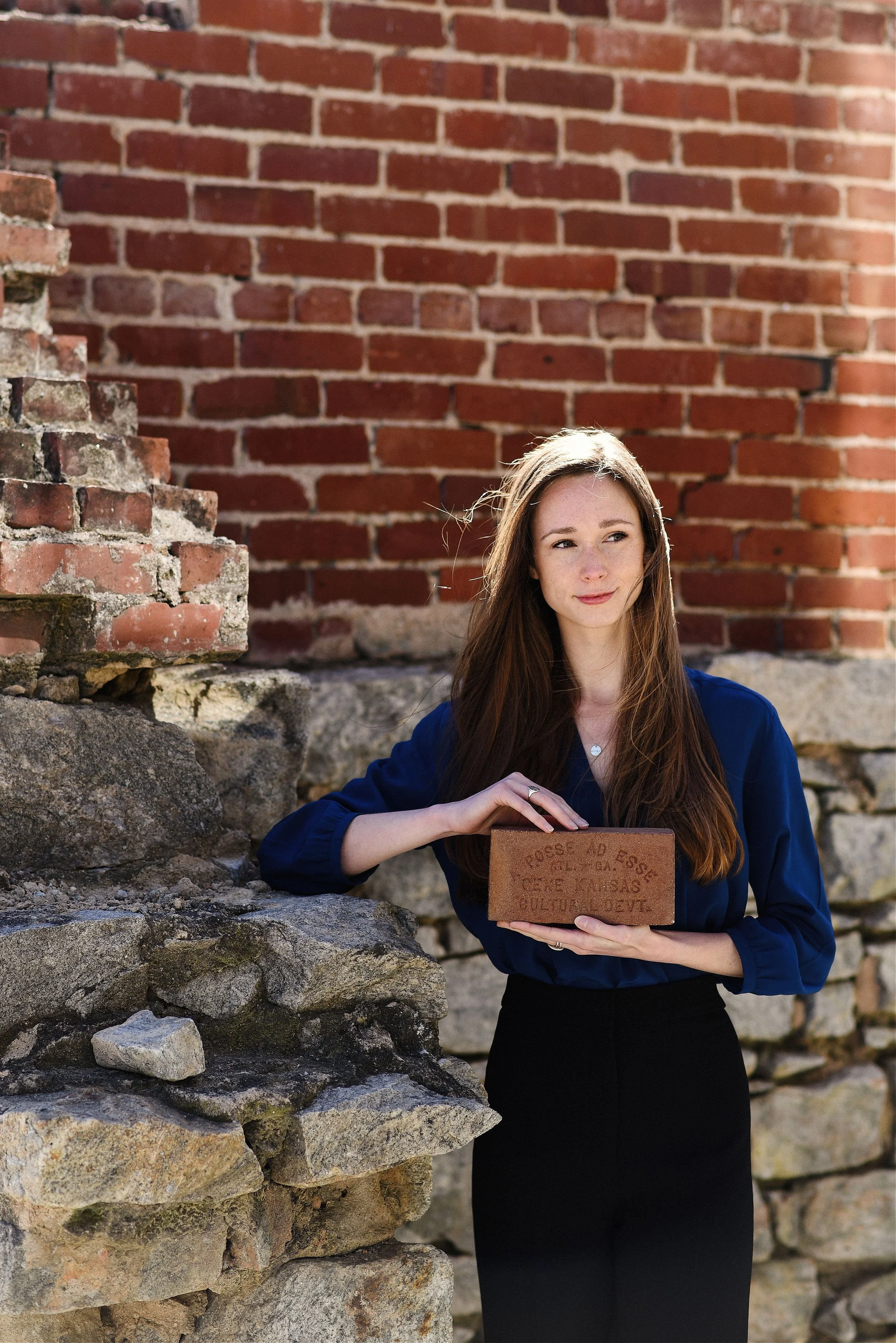 A woman with long brown hair and a blue blouse stands outdoors in front of a brick wall, holding a brick-like plaque with indiscernible text on it.