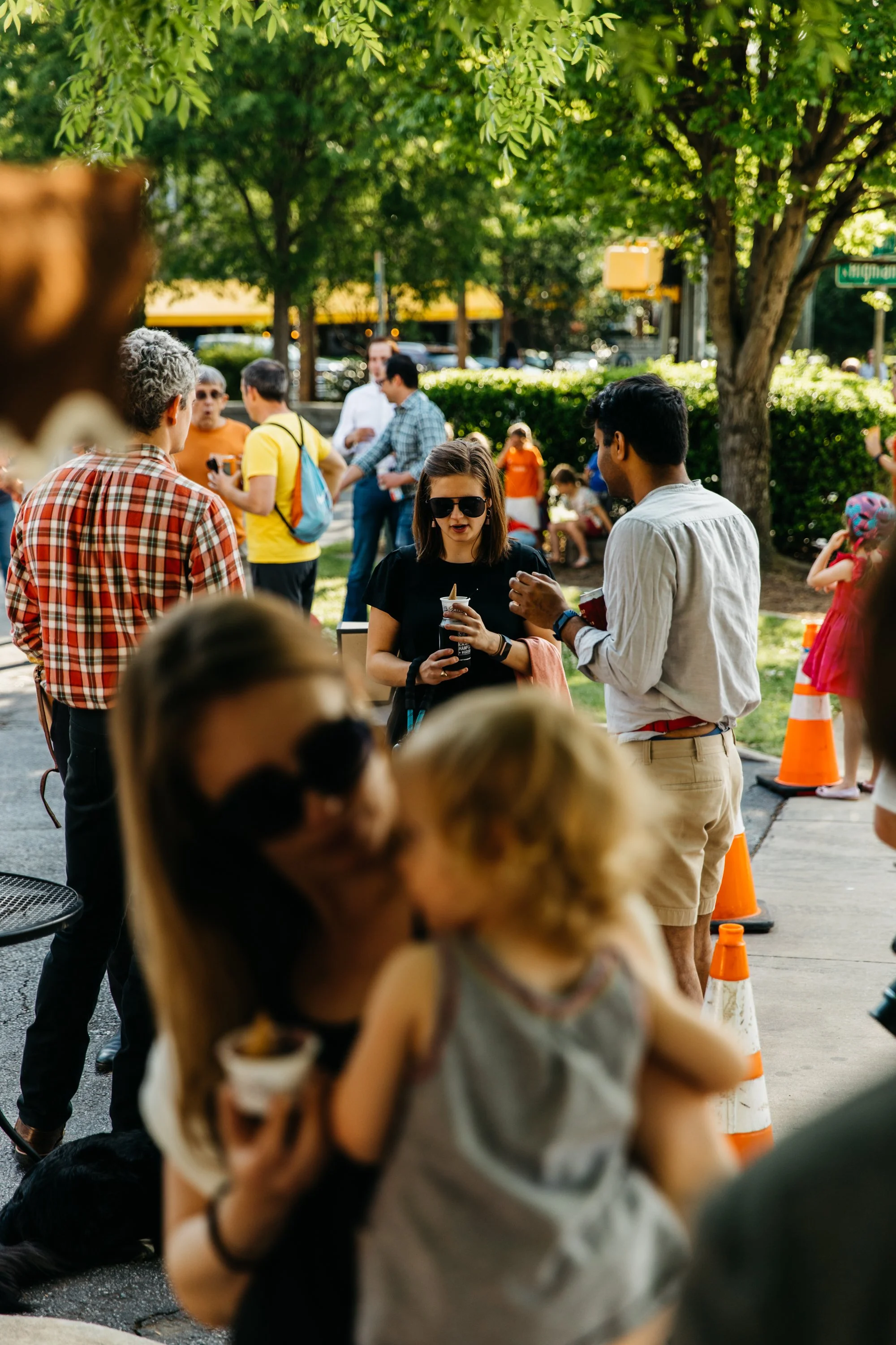 Group of people gathered outdoors on a sunny day, some standing and talking, others sitting near the trees, with orange traffic cones on the sidewalk.
