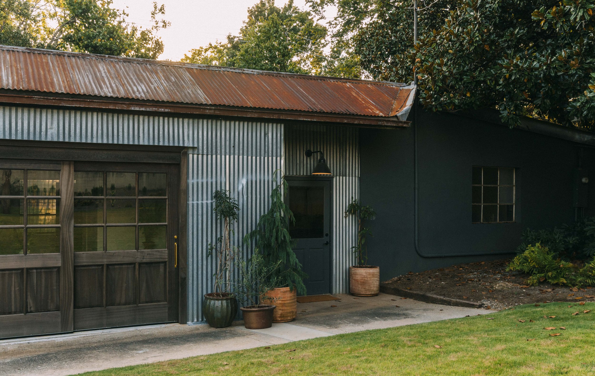 A rustic house exterior with corrugated metal siding and a rusted metal roof, surrounded by potted plants and greenery, with a wooden garage door and a small window, under a partly cloudy sky.