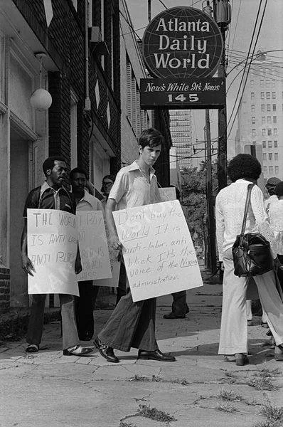 People participating in a protest on a city sidewalk, holding signs about workers' rights, in front of the Atlanta Daily World newspaper building.
