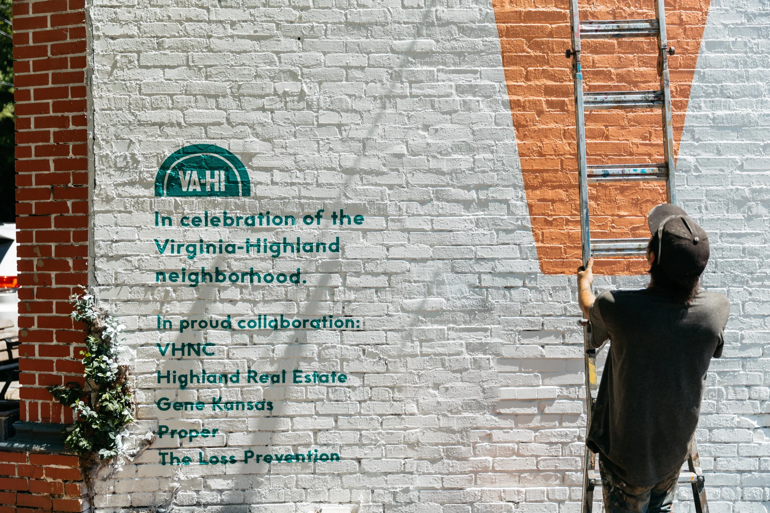 A person on a ladder painting a mural on a brick wall. The mural includes the VA-HI logo and text about celebrating the Virginia-Highland neighborhood and its community partners in Gene Kansas, Kansas City, Missouri.