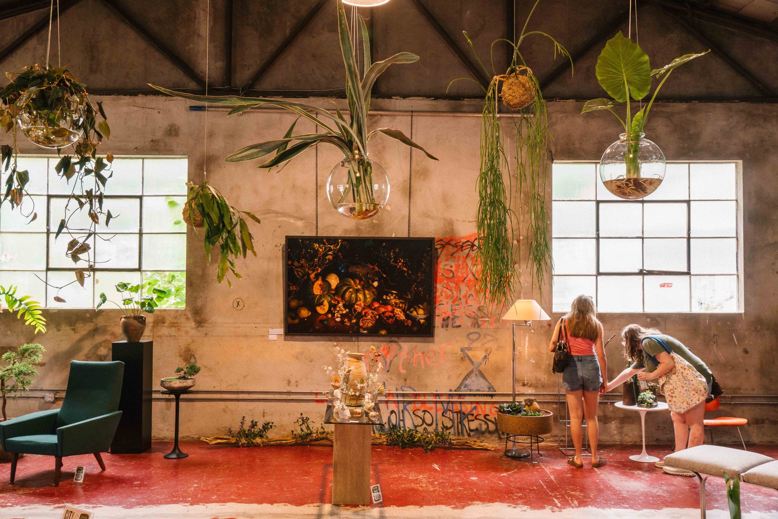 Indoor art gallery or boutique with hanging plants, framed artwork, and two women looking at plants near a white round table.