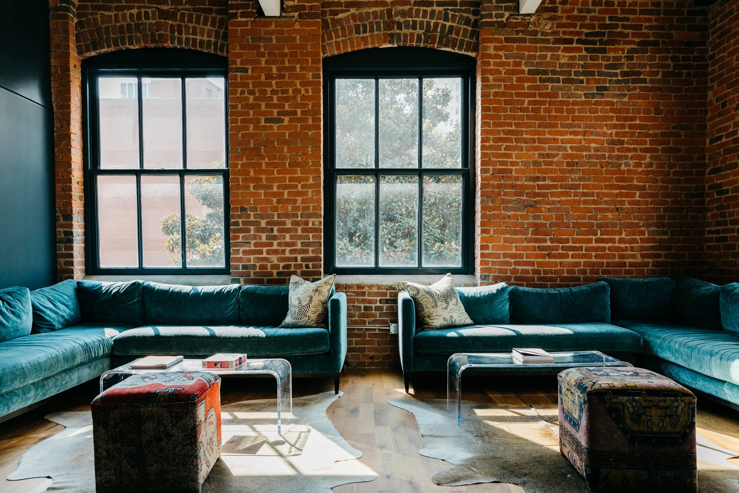 Living room with a brick wall and large black-framed windows, a dark teal sectional sofa with patterned pillows, a glass coffee table, and colorful upholstered ottomans.