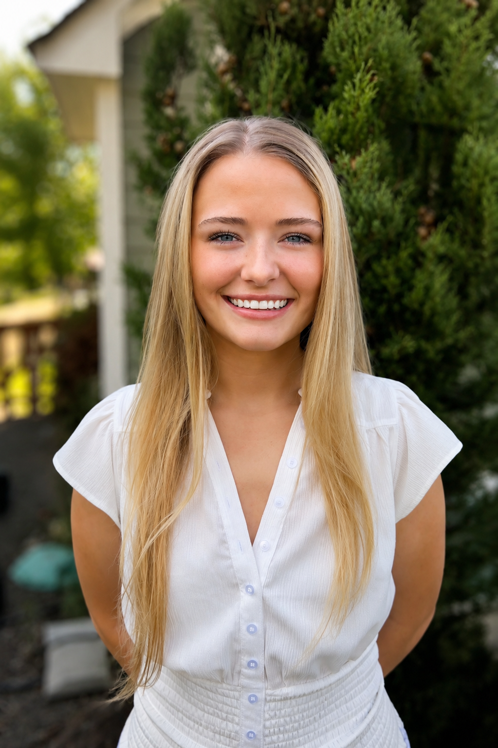 A young woman with long blonde hair and blue eyes smiling outdoors, wearing a white blouse in front of green bushes and a house.