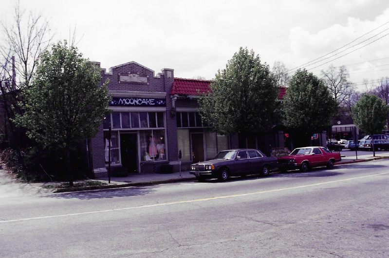 Street view of a small retail building with a sign labeled 'MOONCAKE', parked cars, and trees in front of the sidewalk.