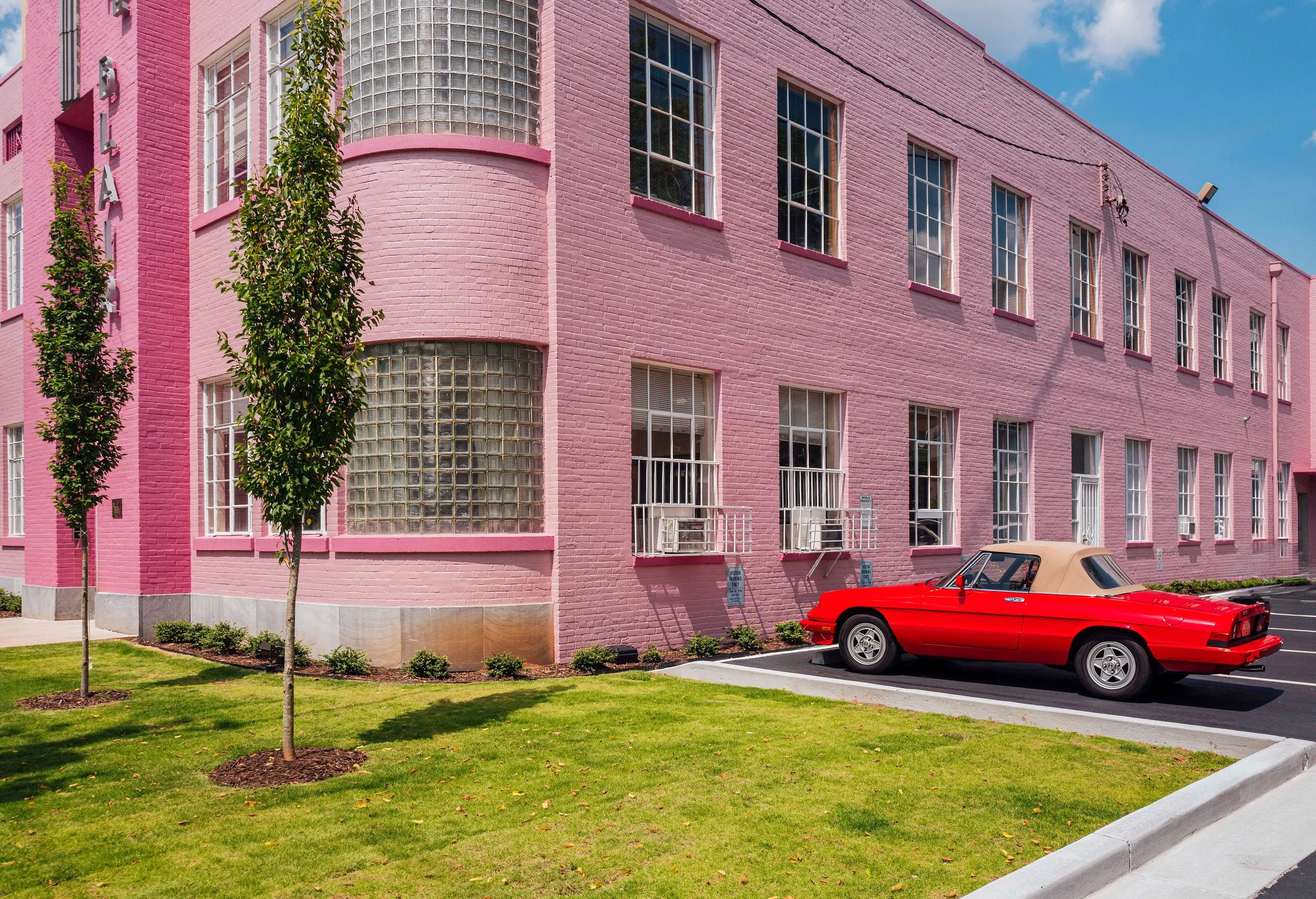 A pink brick building with multiple tall windows, some with small balconies. There are two young trees planted in front of the building, a grassy area, and a red vintage convertible car parked in the parking lot.